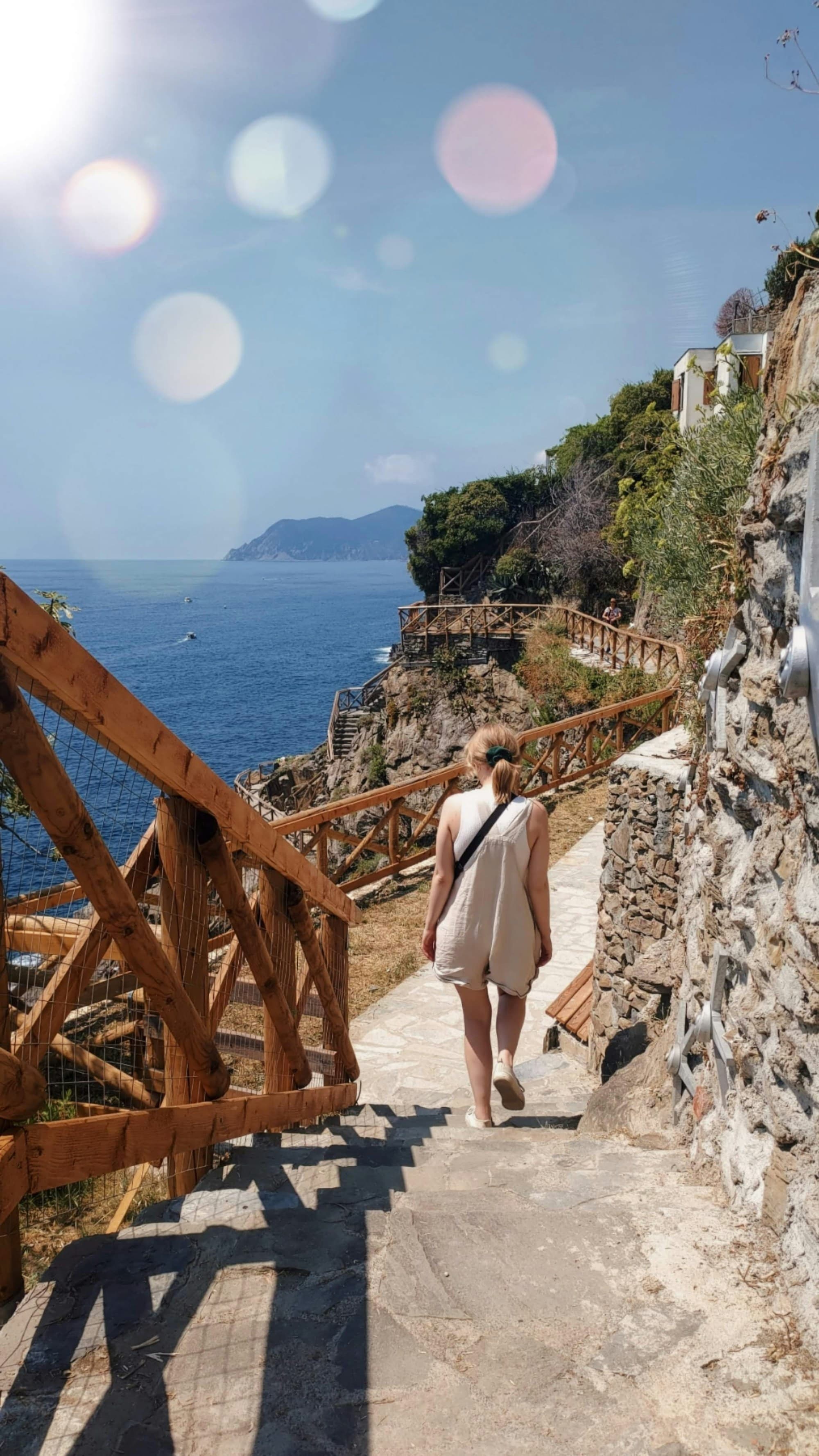 The image captures a person strolling along a coastal pathway, with the sea and mountains gracing the horizon.
