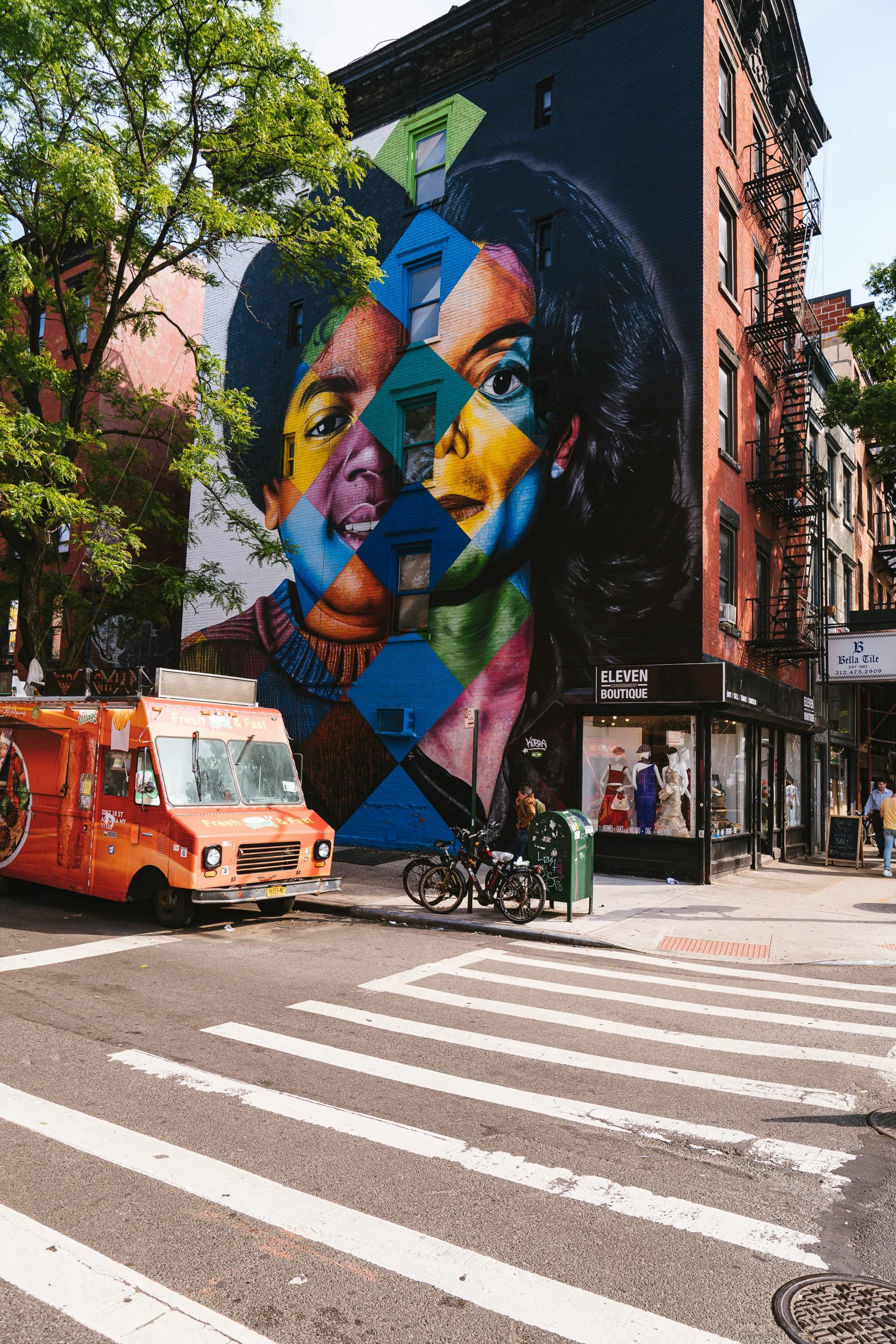 The image depicts a lively street scene with a striking, colorful mural of a face painted on a building’s side.