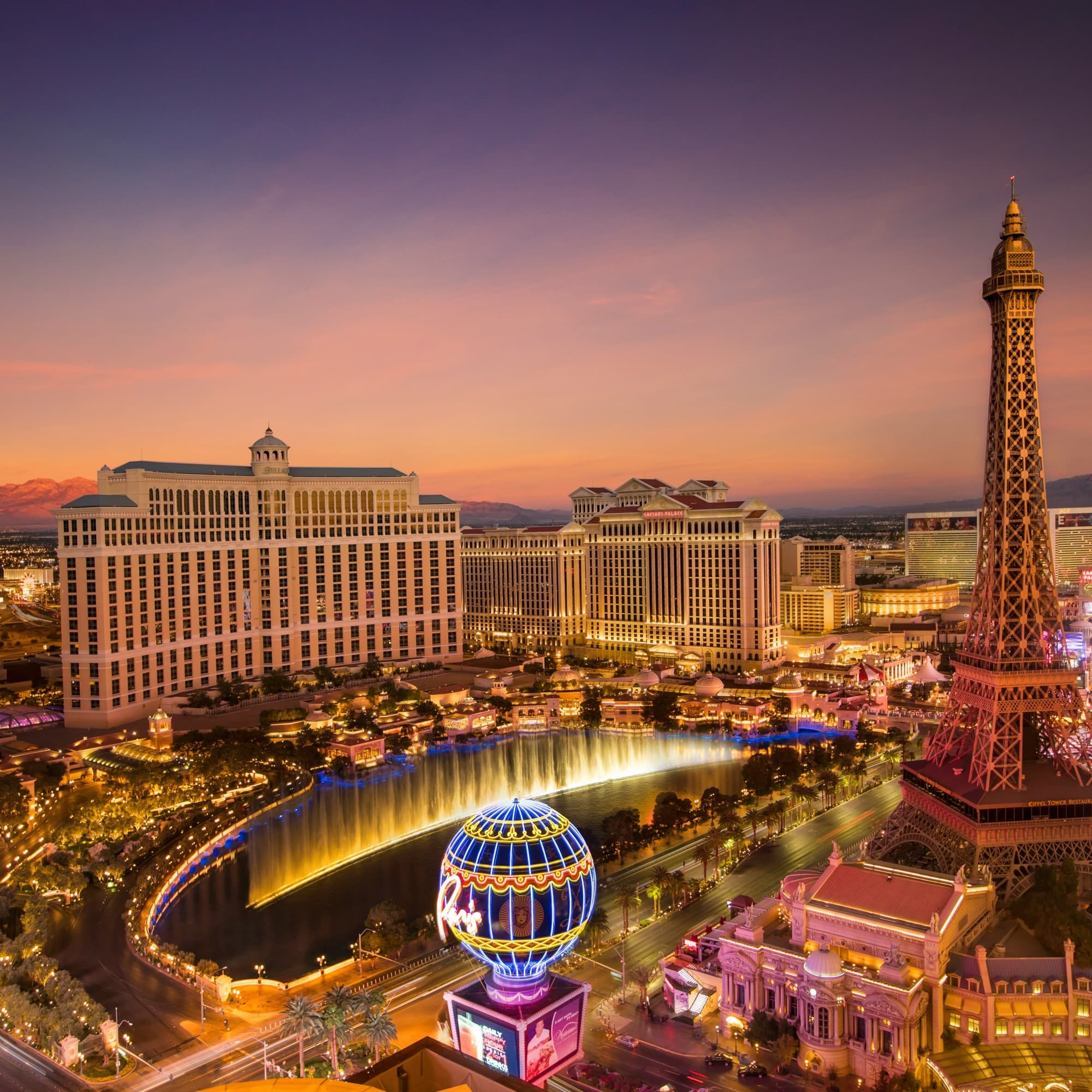 An aerial view of the Bellagio hotel and fountain in Las Vegas, with the mock Eiffel Tower, and city lights underneath a dusk sky.