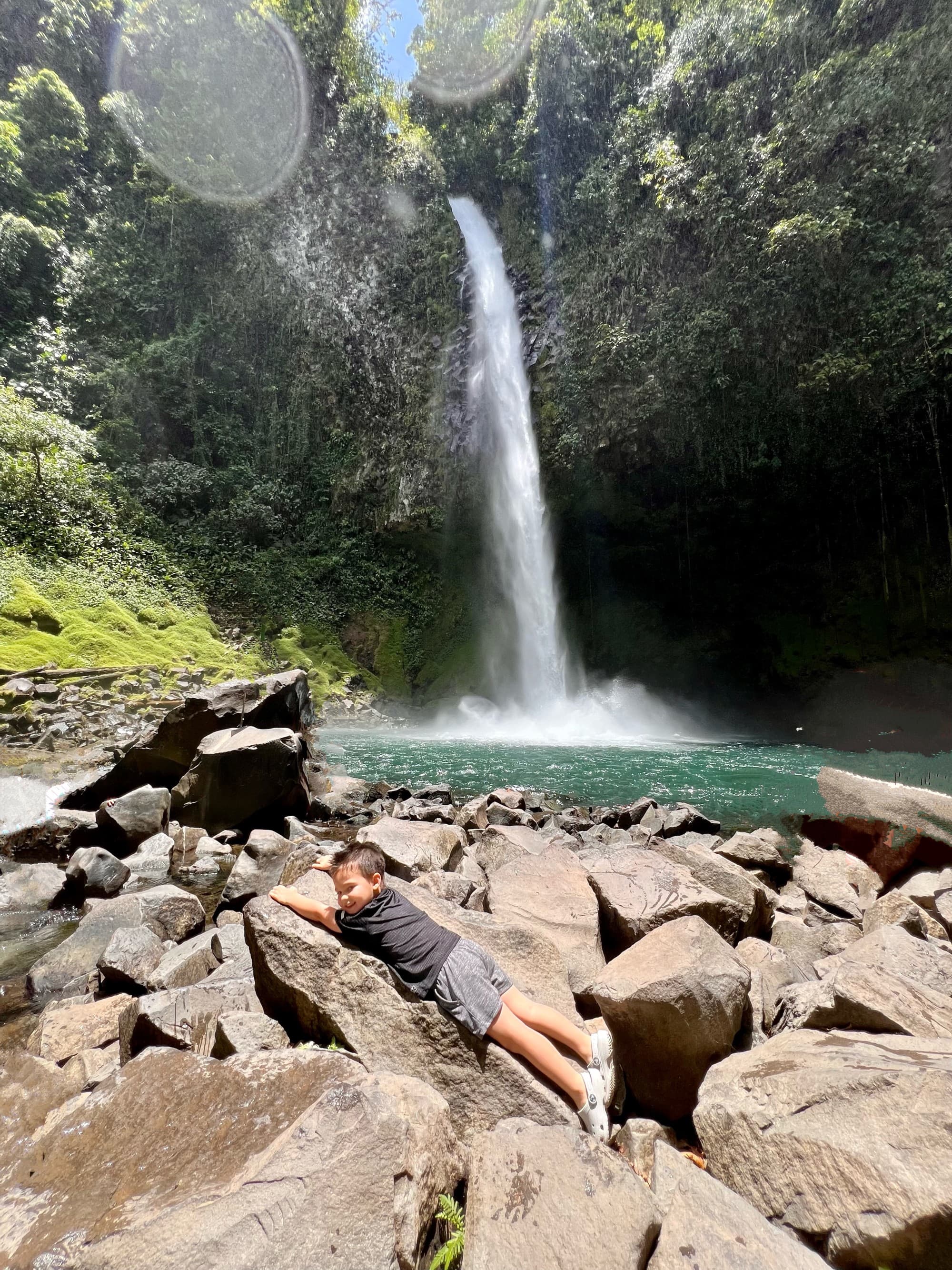 A tranquil scene with a child resting on some rocks by a waterfall amid lush greenery.