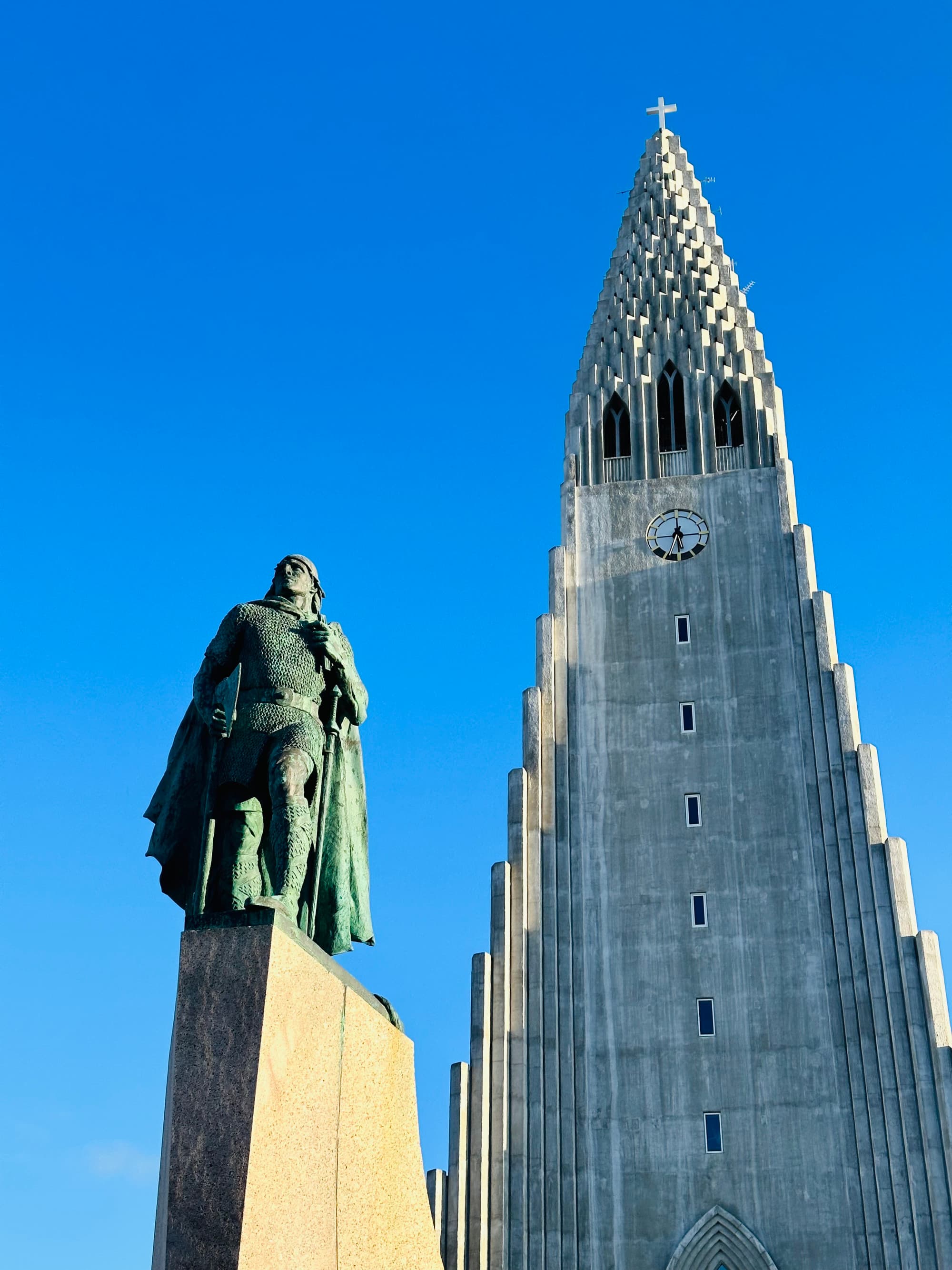 The image portrays a statue in front of a towering, pointed church against a backdrop of clear blue skies.