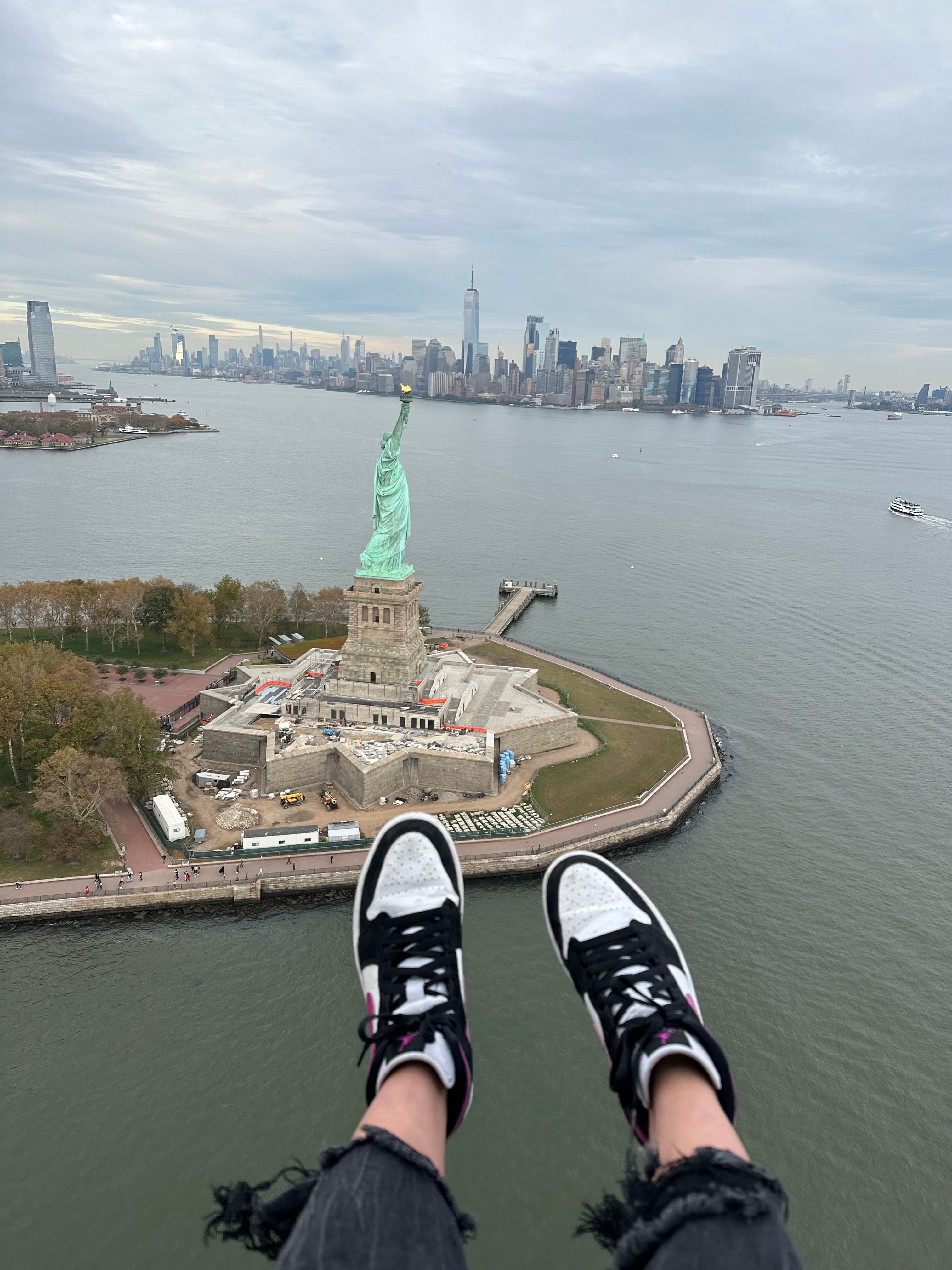Travel Advisor's feet hanging off of an elevated ledge over the Hudson River, with Lady Liberty and New York City's skyline in the background.