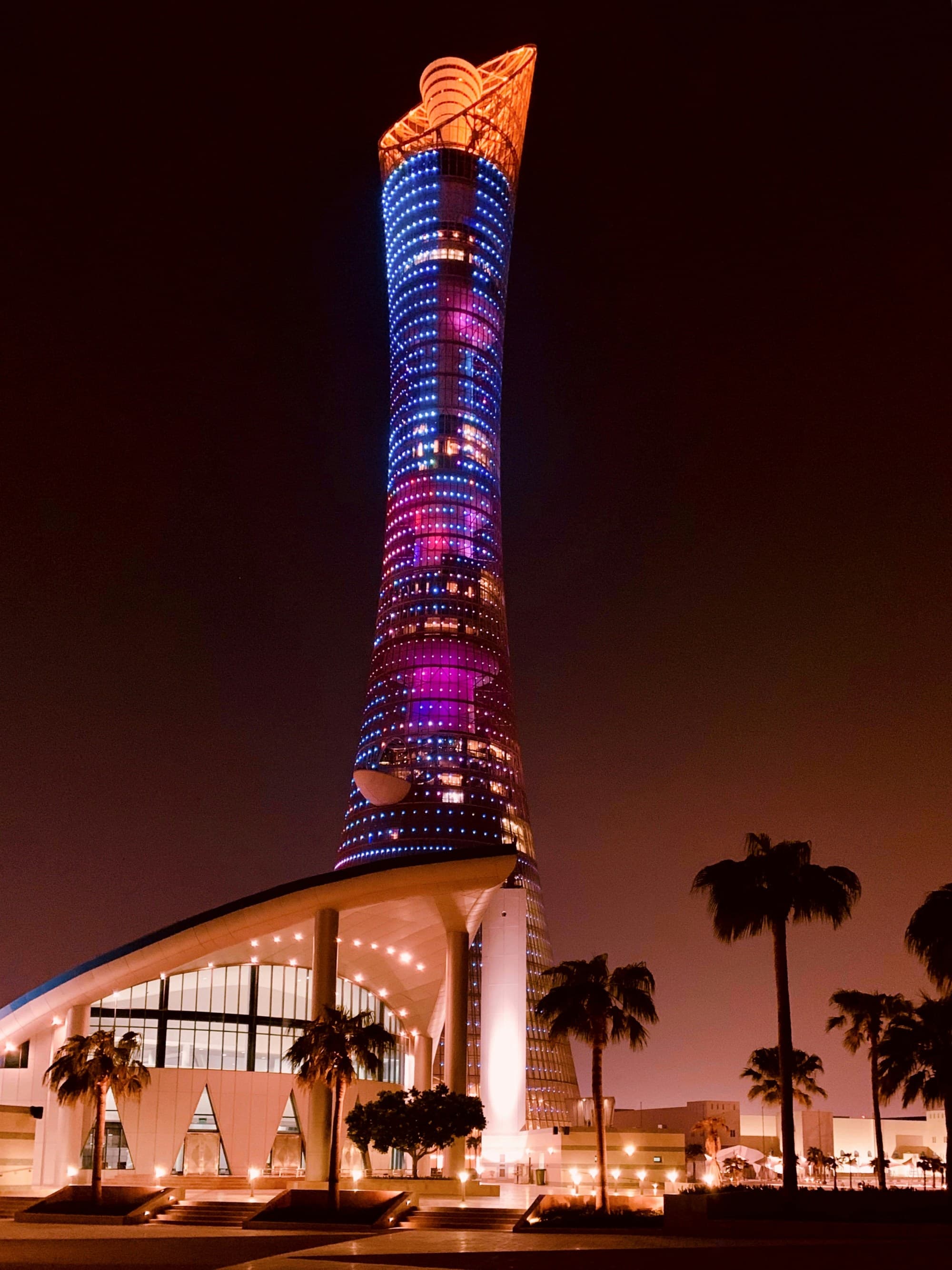 A modern skyscraper illuminated with blue and purple lights stands tall against the night sky, flanked by palm trees.