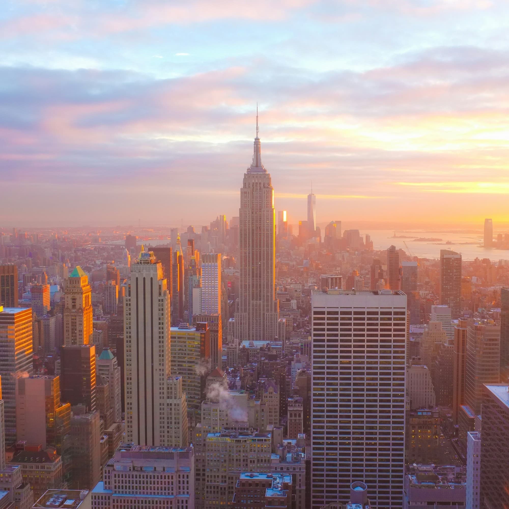 A view of Midtown Manhattan's skyline, the Empire State Building, Hudson River and Lady Liberty underneath a colorful sunset.