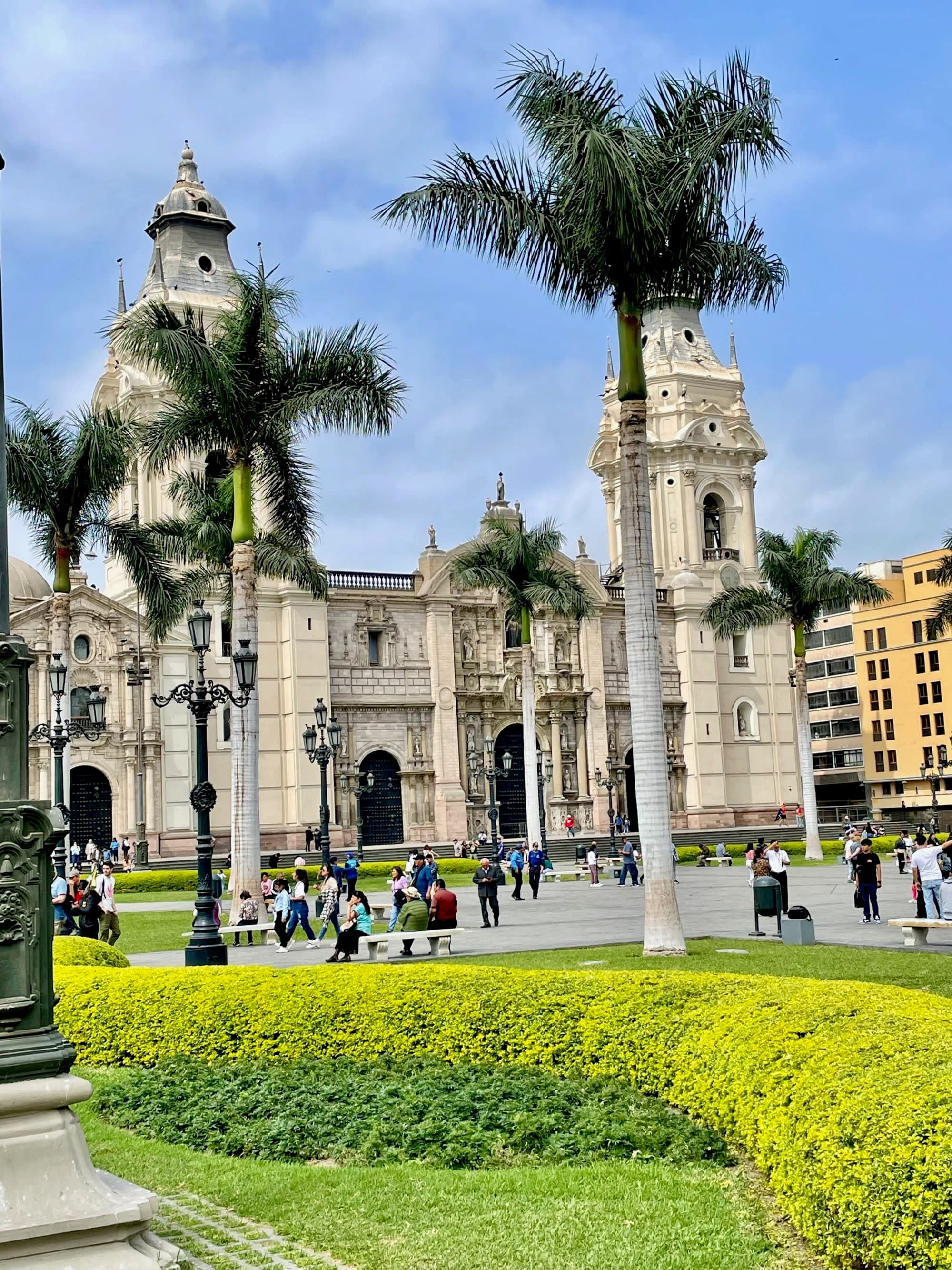 This image depicts a vibrant city square with a stone building, palm trees, a green hedge and people walking around in the surrounding areas.