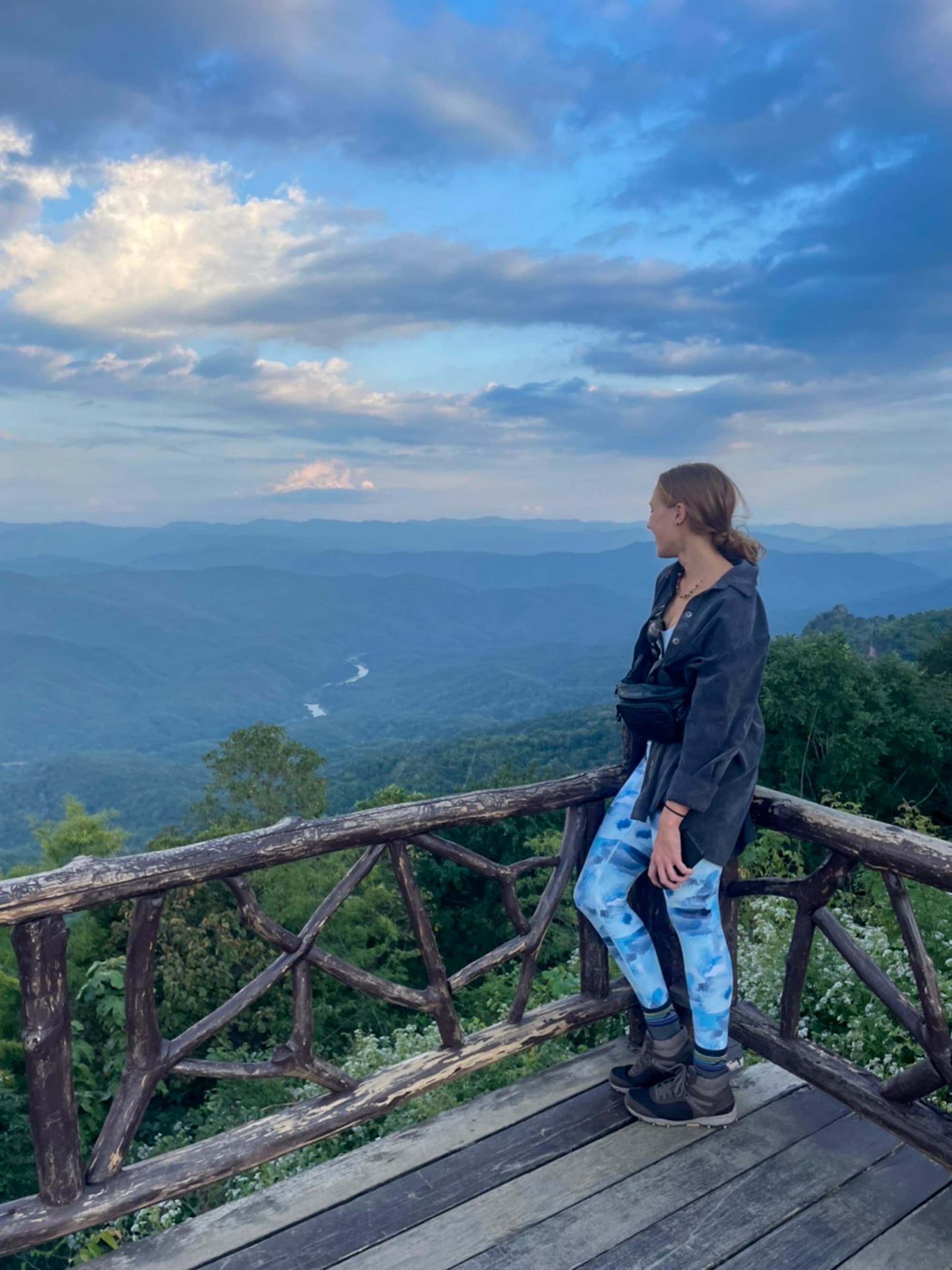Advisor on a wooden platform overlooking a scenic mountain vista, with a winding river or road below and a partly cloudy sky above.