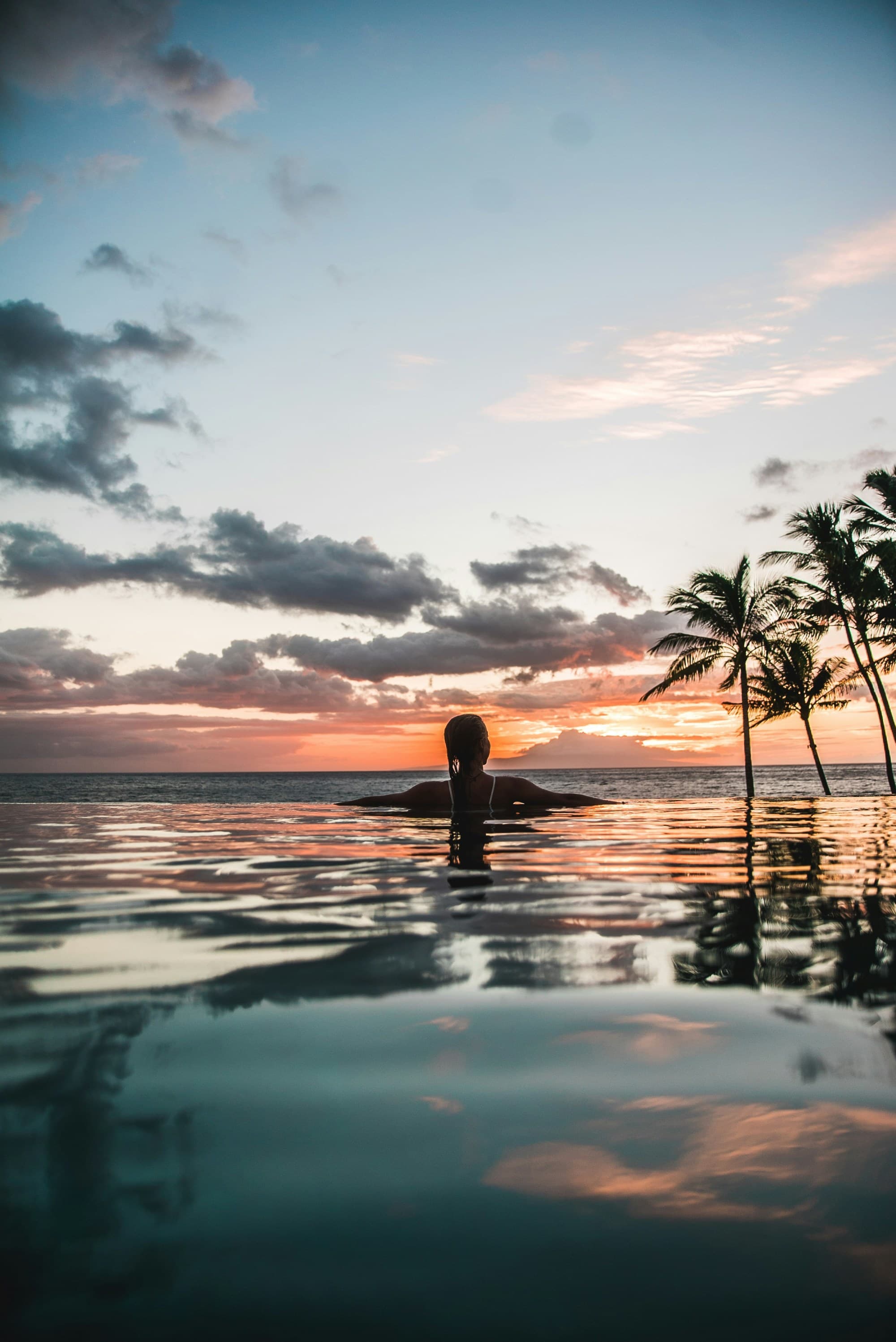 A serene sunset scene with a person immersed in water, silhouetted against a sky ablaze with vibrant sunset hues and palm trees.