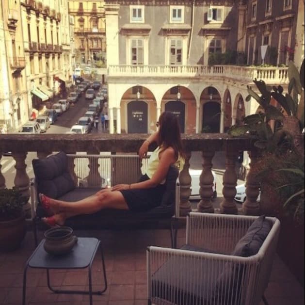 Wieteke sitting on a balcony overlooking a Sicilian street, surrounded by plants and classical architecture.