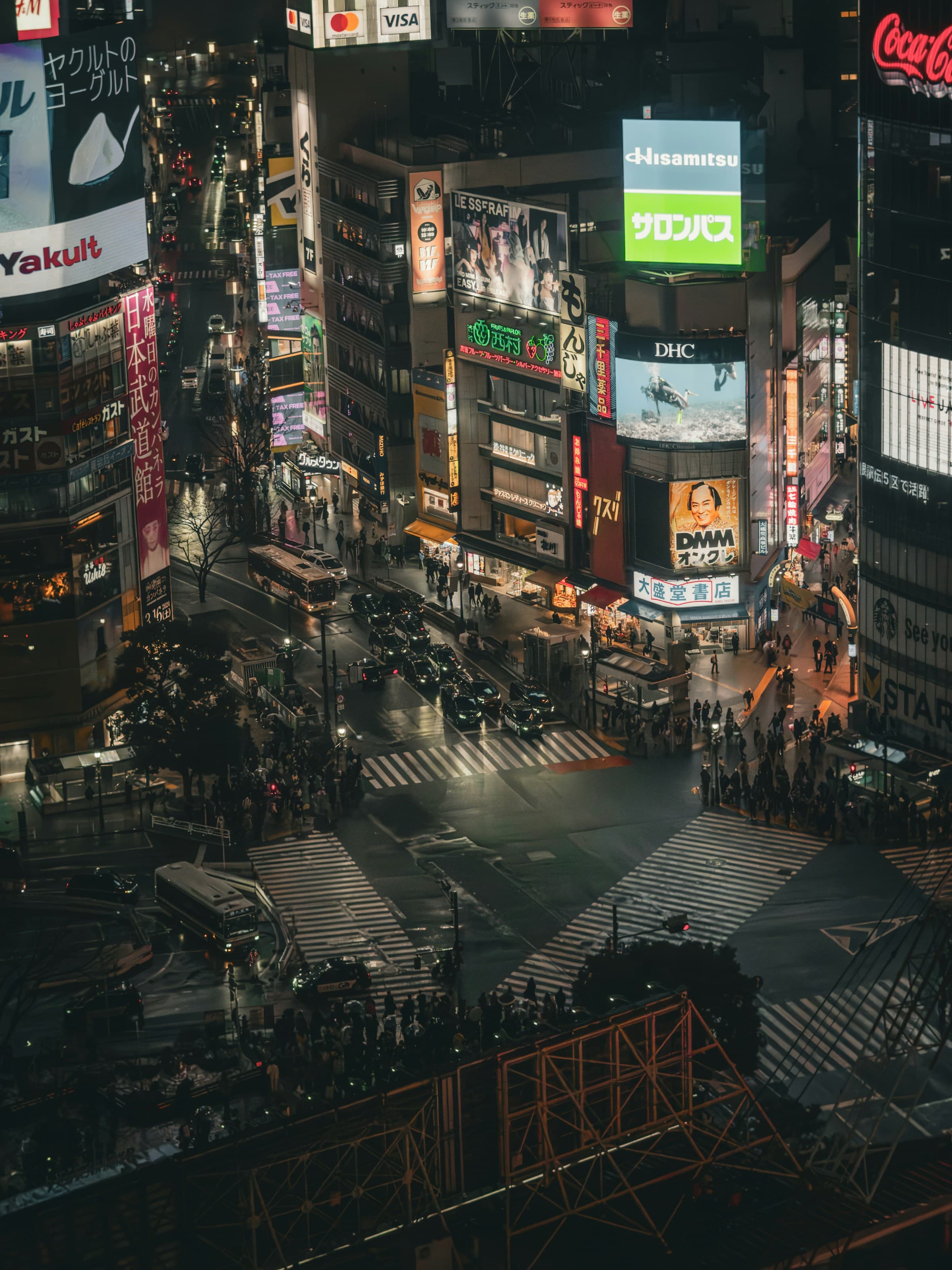 An image of an overview of Tokyo's city center with various lights, billboards and LED screens. There are people walking along the street down below.