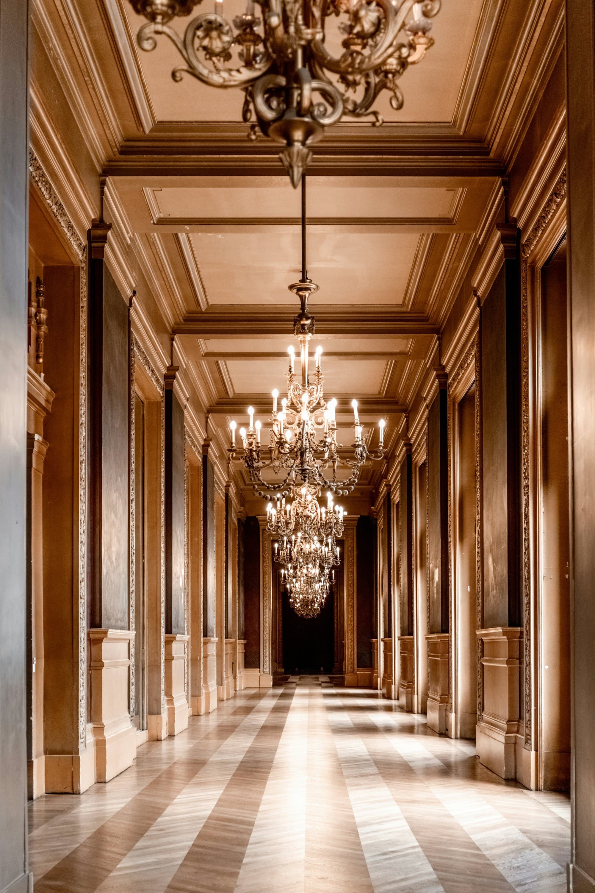 Grand chandeliers hanging from a ceiling in a hallway with striped tile flooring and detailed paneling.