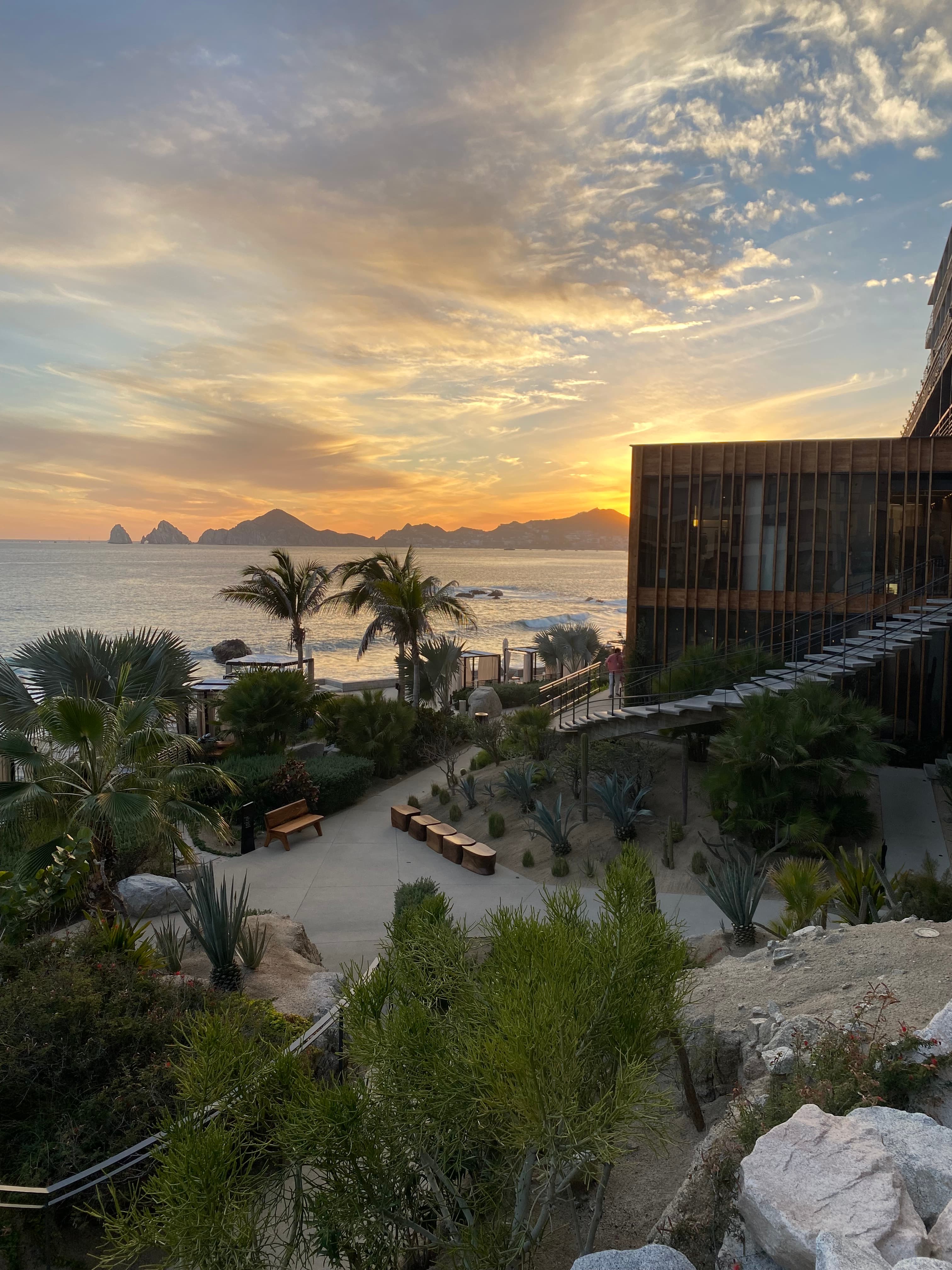 A picture of a sunset shining over mountains, water and a hotel resort with trees, paths and bushes.