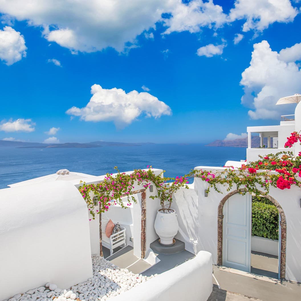 A picture of a white terrace with pink and green flowers, a white pot, an archway and a cloudy blue sky above with the sea in the background.