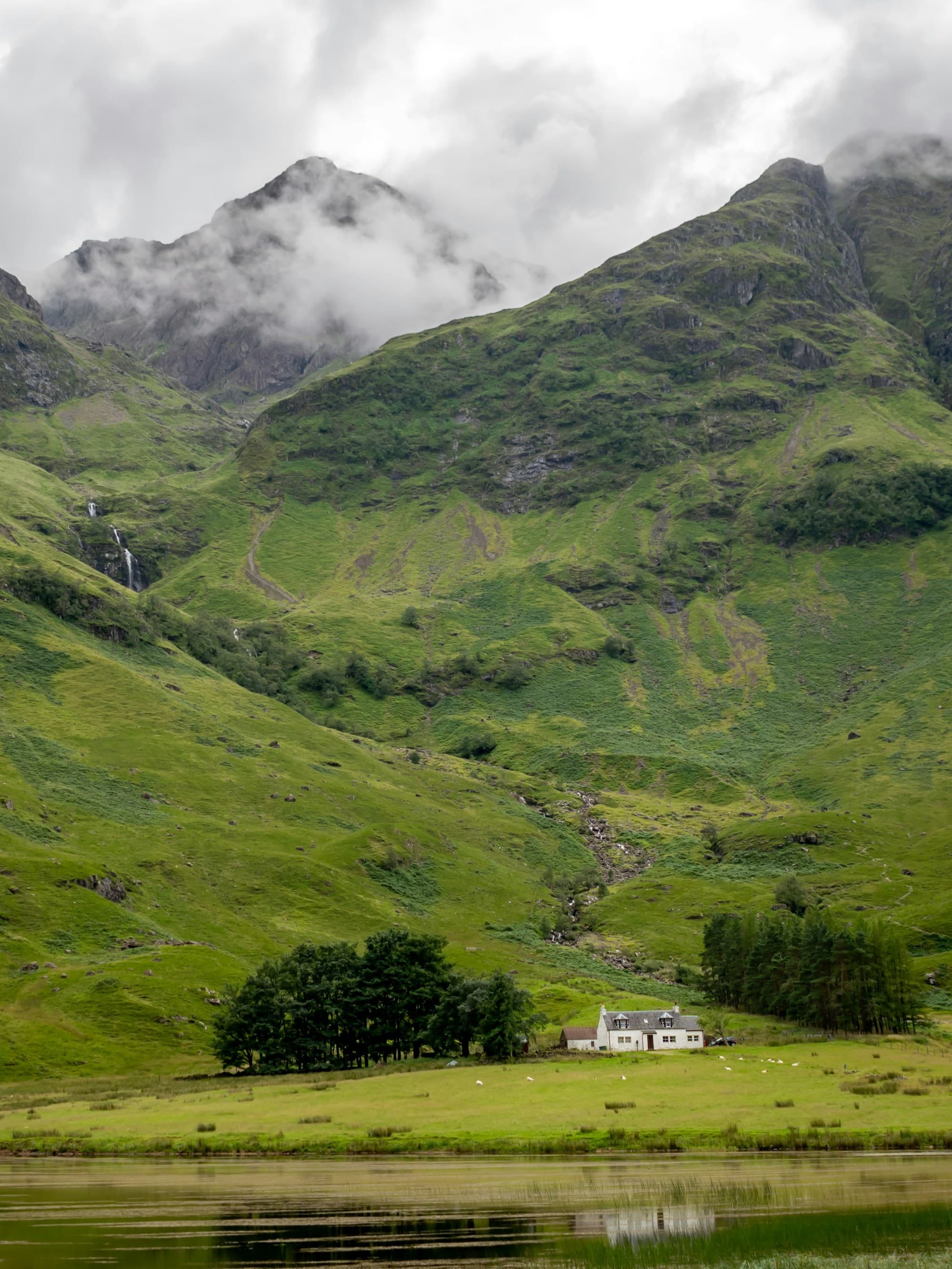 A view of a small house nestled into the base of a large and bright green mountain with fog surrounding the peaks.