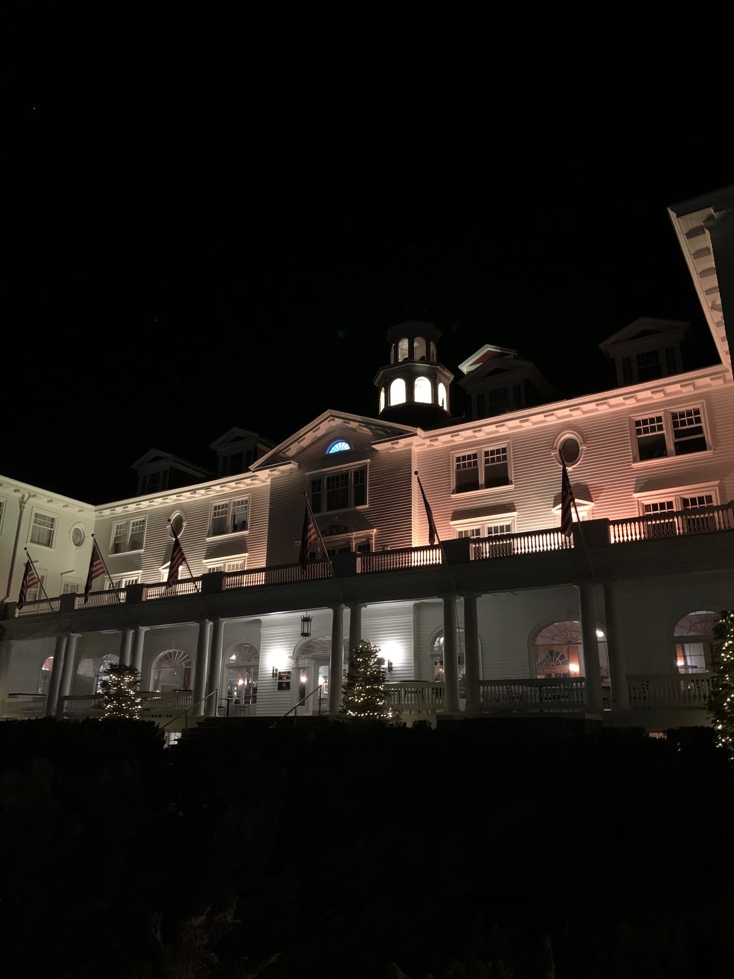The exterior of Stanley hotel illuminated at night with eerie lighting and US flags flying and christmas lights.