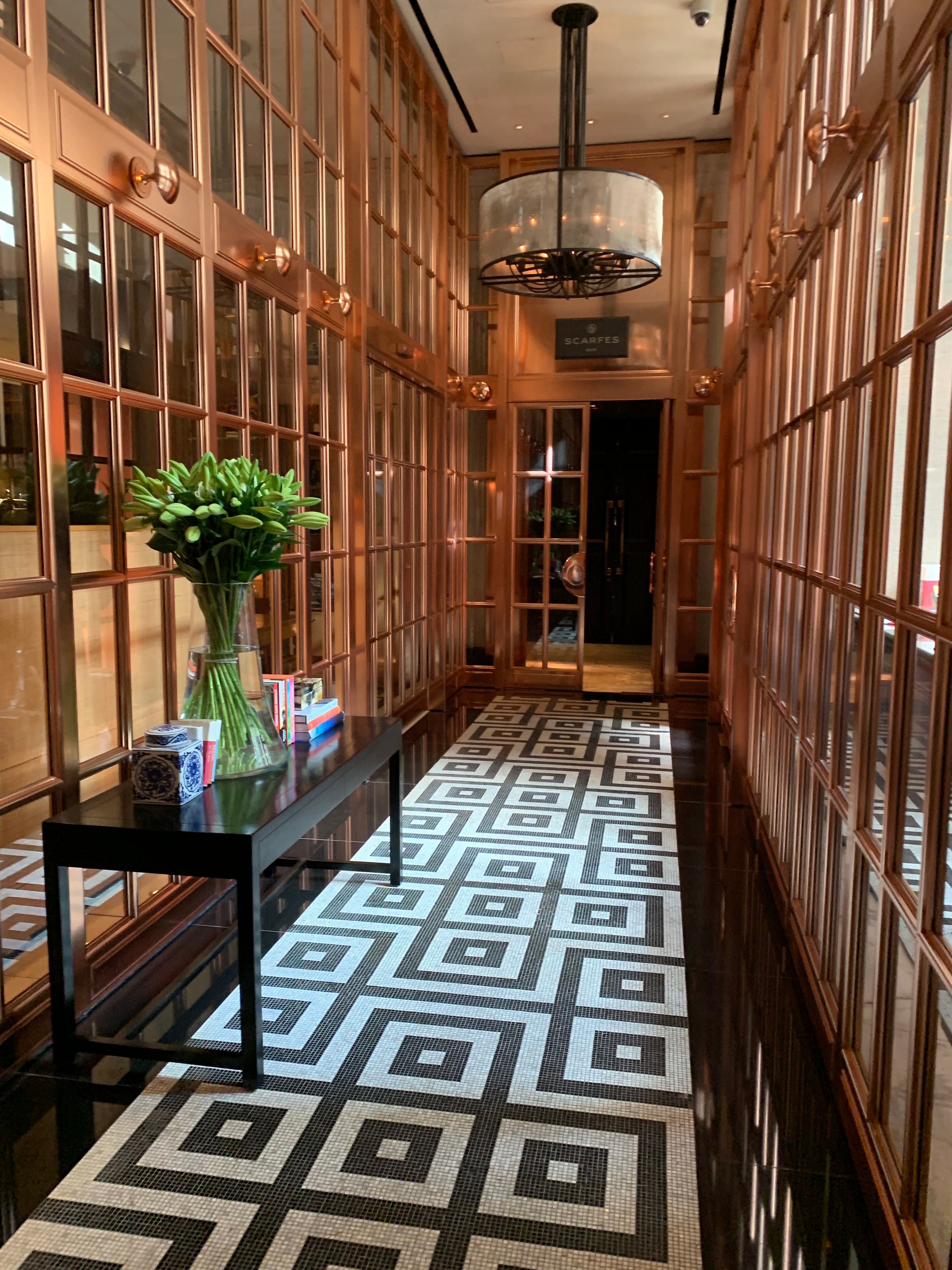 A swanky hallway of a London hotel, with square-patterned tiles, and glass walls with wooden panes