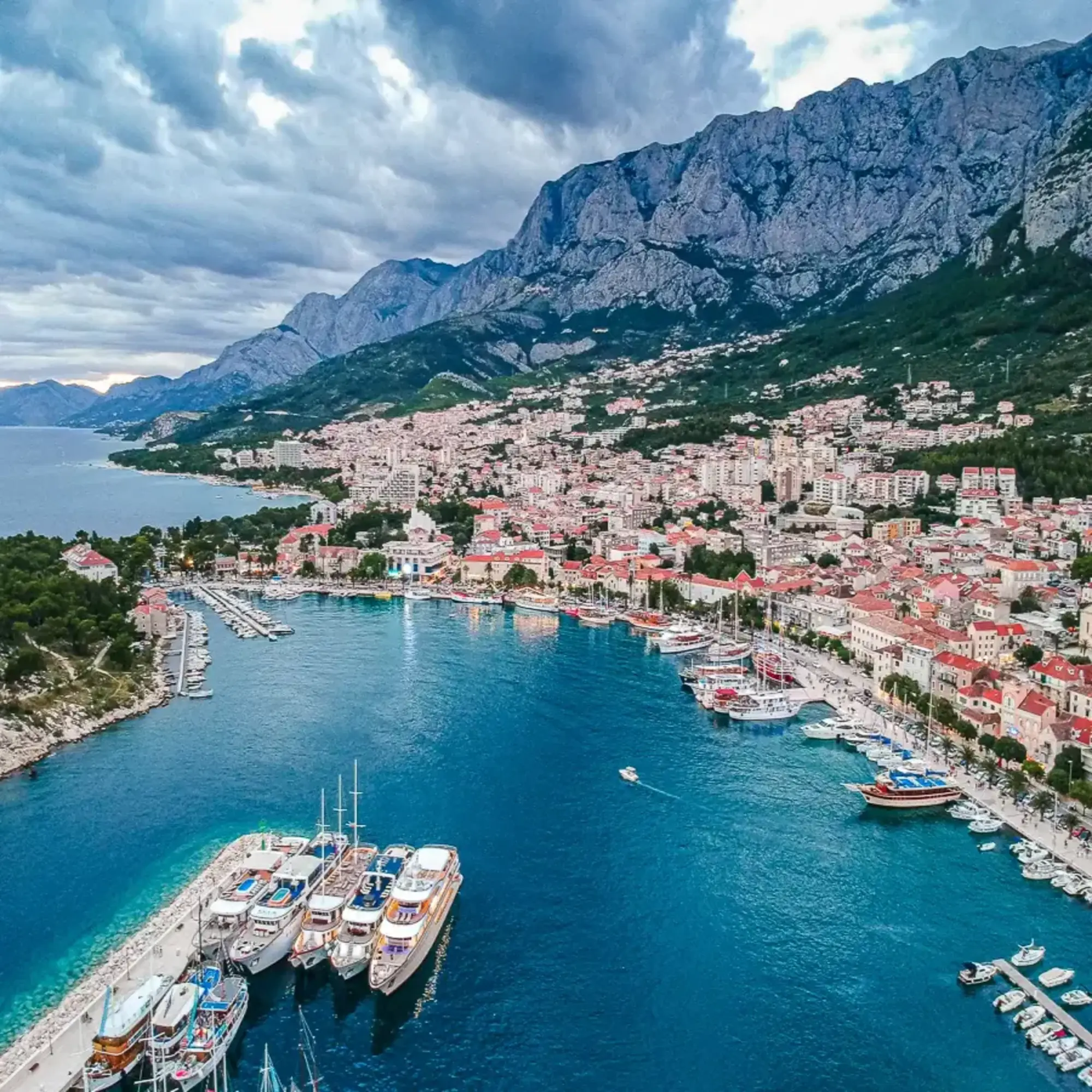 A view of the harbor, anchored boats and city of Croatia complete with red-roofed buildings, rocky mountains and trees.
