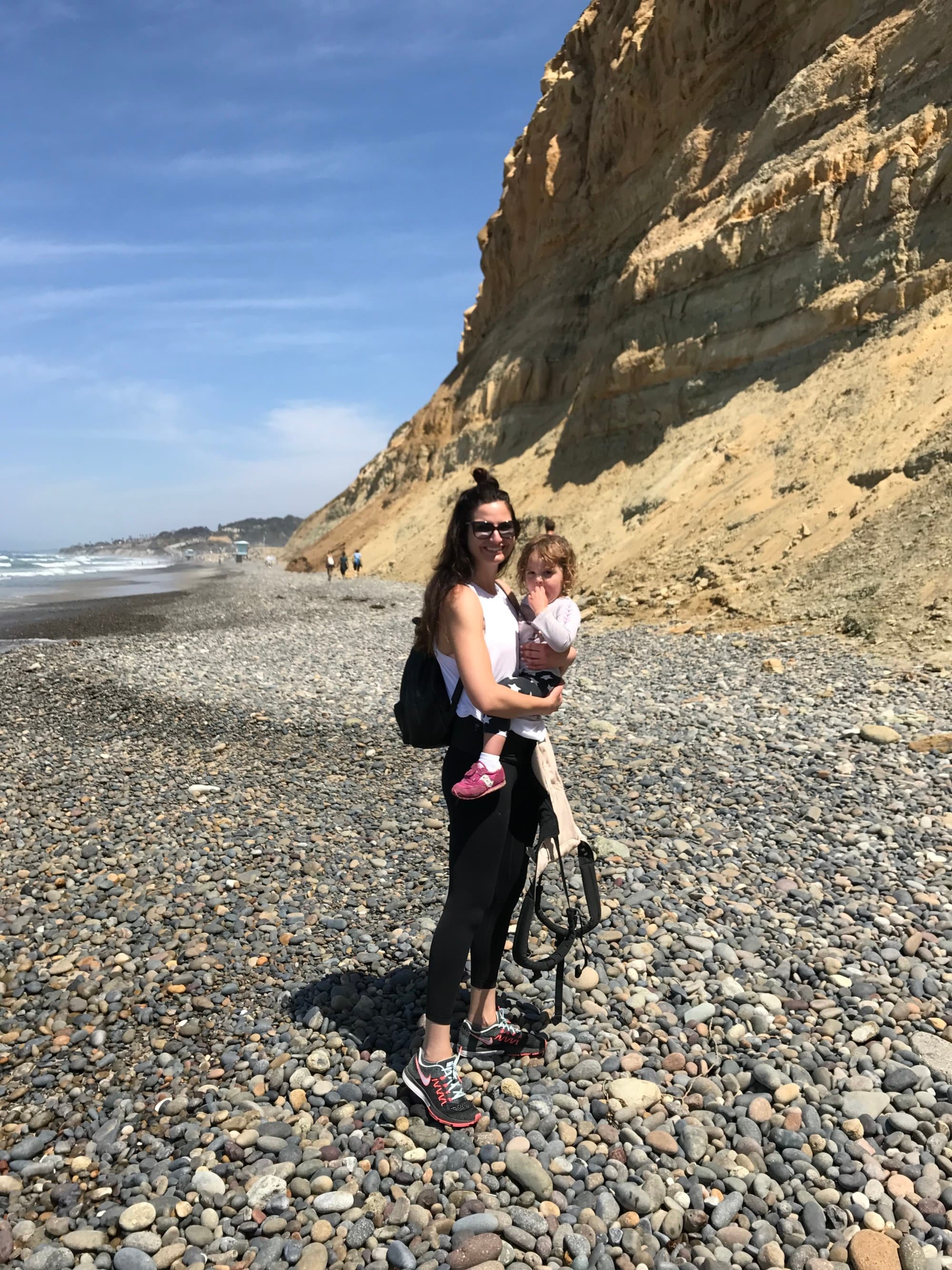 A picture of a woman holding a baby near a beige mountain on top of a rocky beach at daytime.