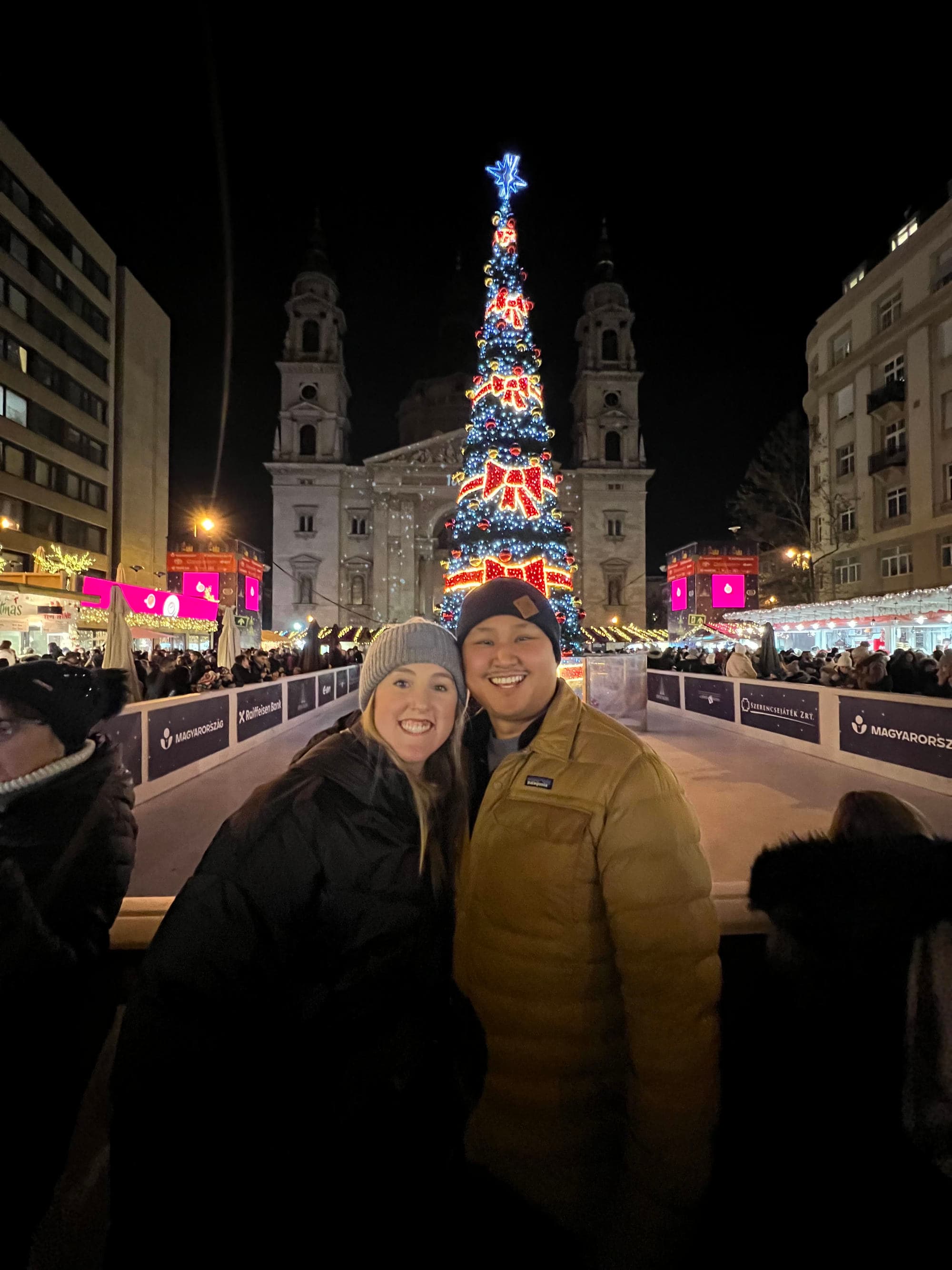 A couple posing for a photo in front of a lit up Christmas tree in a city square at night.