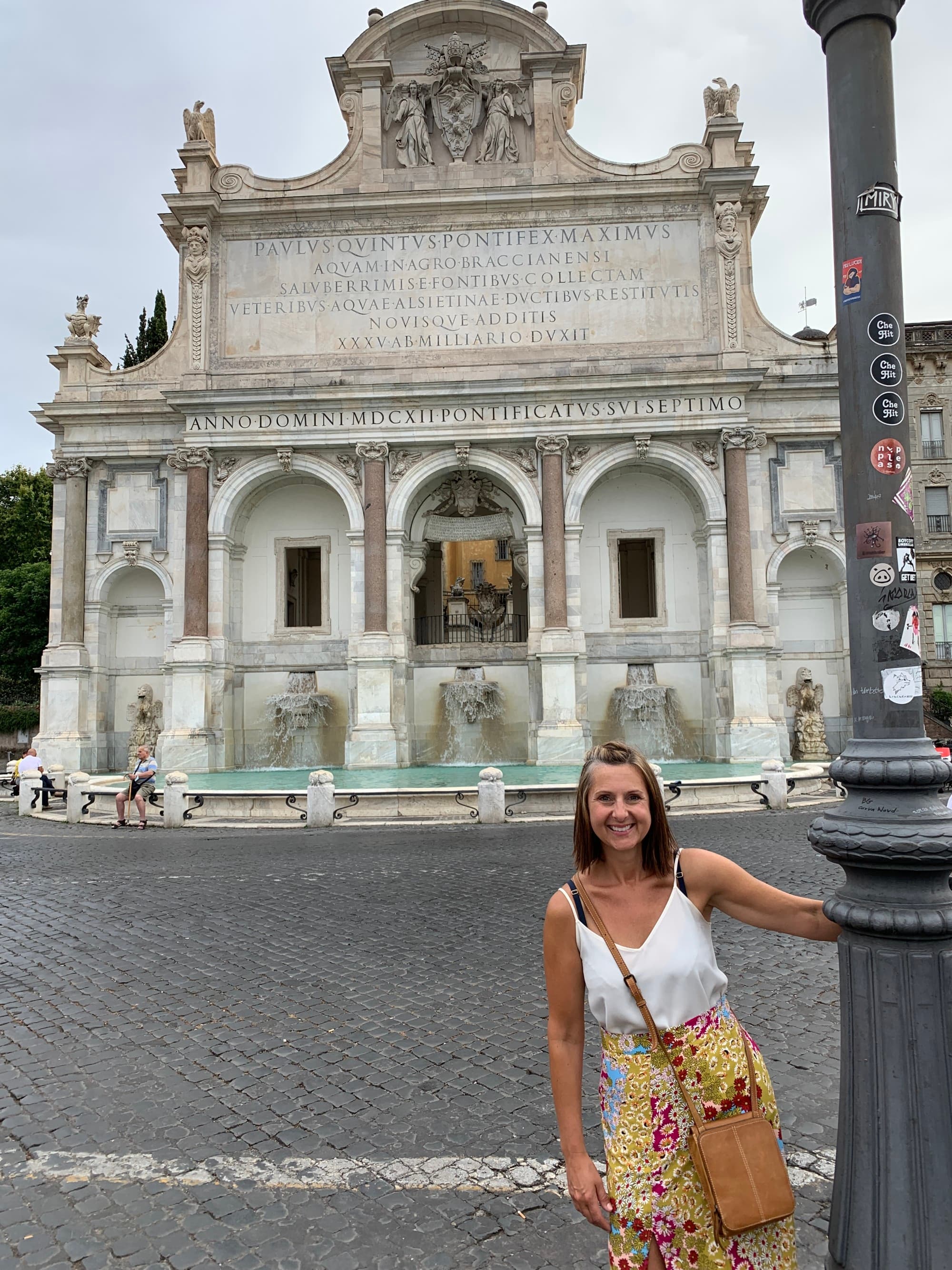 A women in white top standing in front of an old building with statues and text written on it.