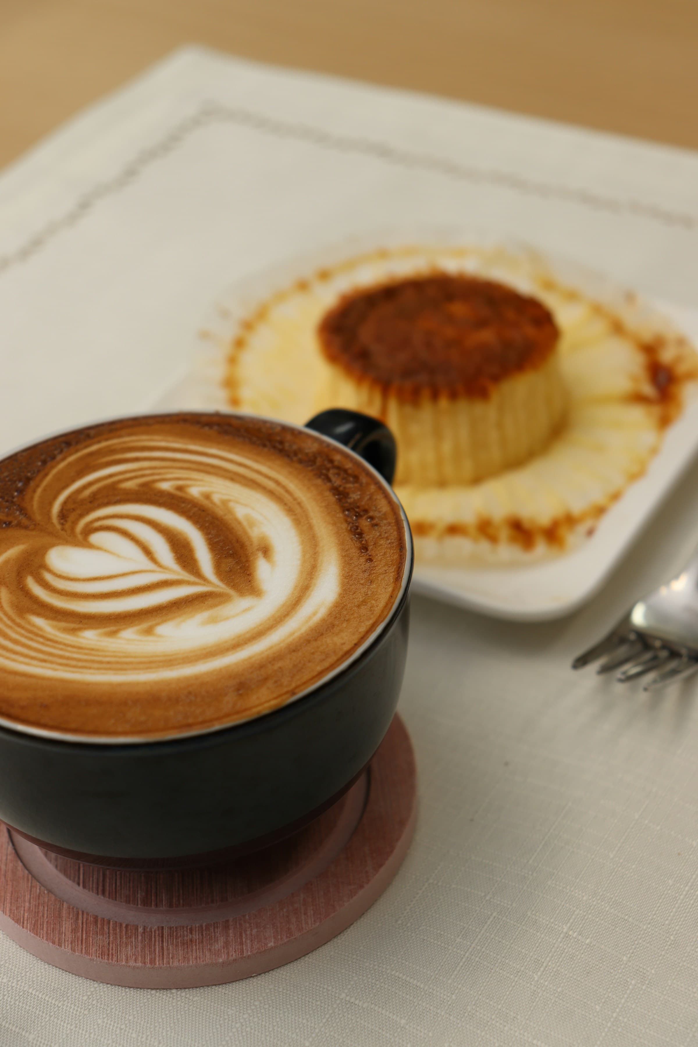 Coffee latte art (wing heart) and cheesecake on a plate sitting on top of a table.