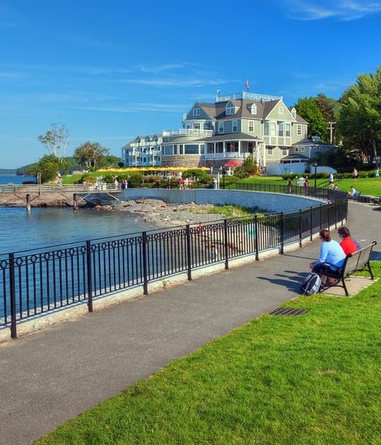 A view of a path, a bench with two people sitting on it, a grassy lawn and a harbor with a house in the corner right side of the image.