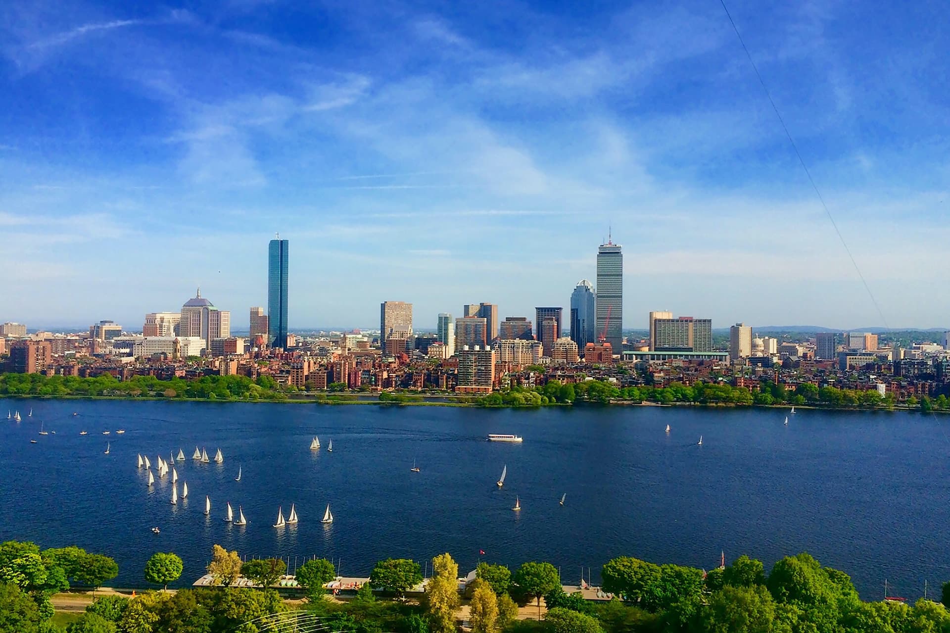 A beautiful view of the Boston skyline with the vibrant blue harbor in the forefront. There are boats and trees surrounding the skyscrapers, water and industrial views.