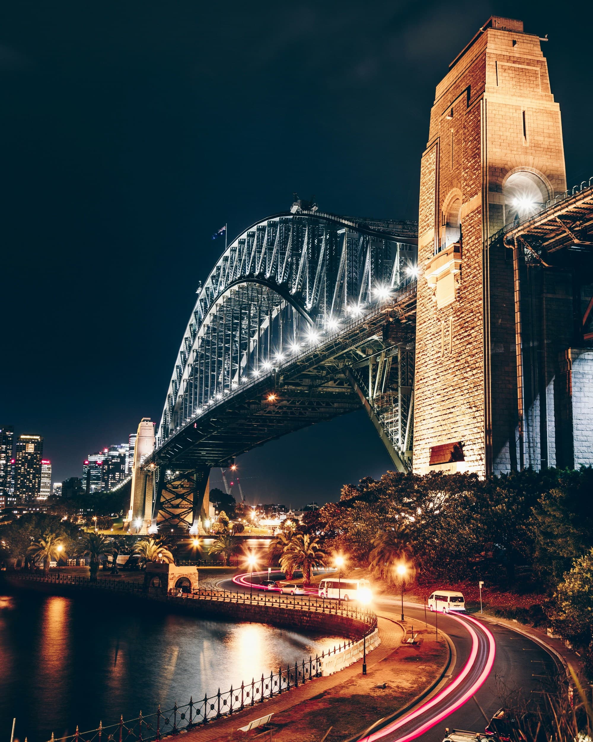 A view of a road, body of water and large arched bridge at nighttime lit up with lights.