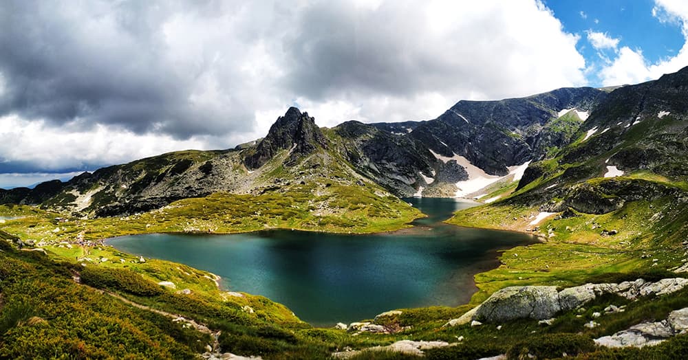 Sofia Bulgaria green grass with mountains, snow, blue lake and low white clouds and blue sky