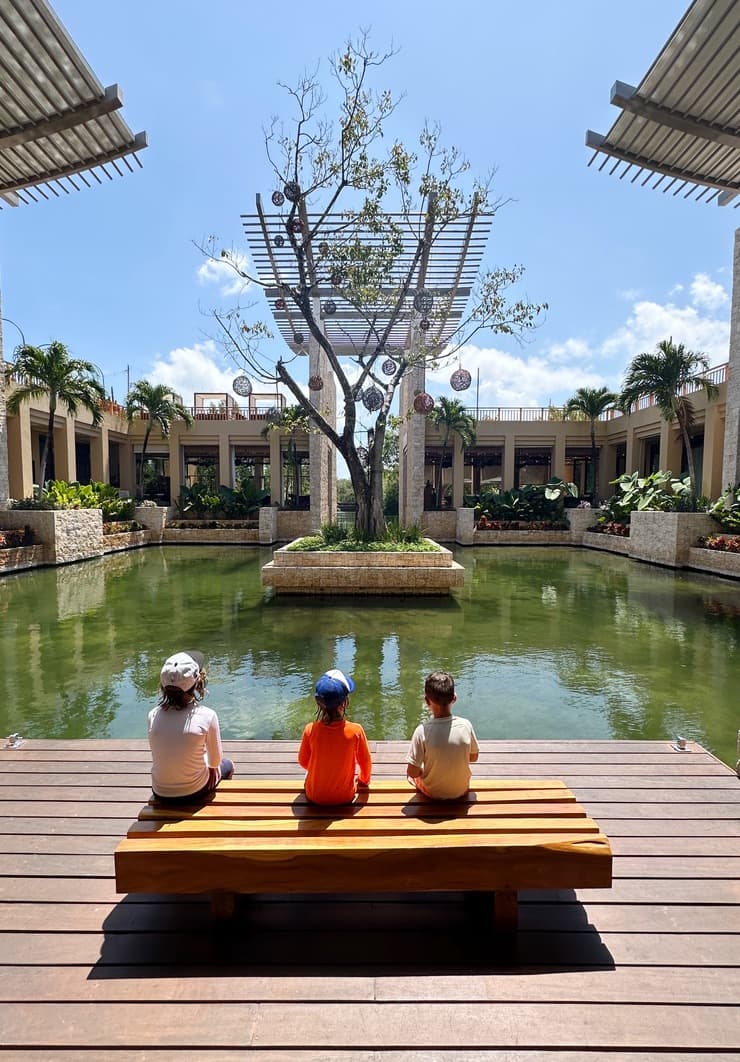 Three kids sitting on a wooden bench next to a pond on a sunny day.