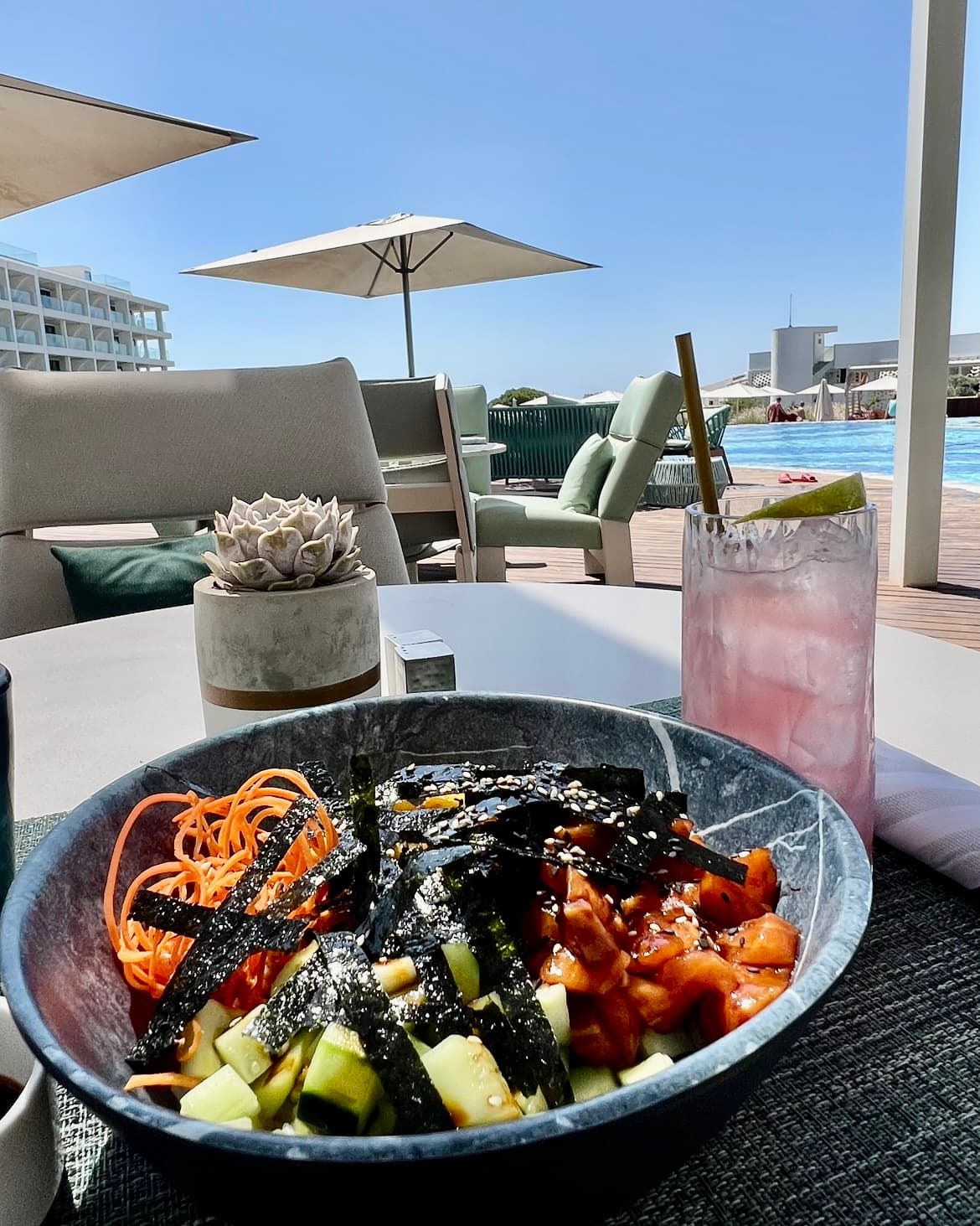 A colorful bowl of food with a pink cocktail on a table in the pool area.