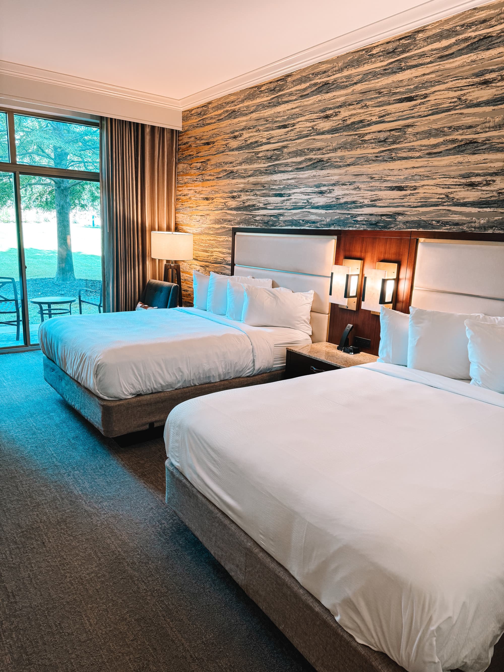 A double queen room features two spacious beds and a view of the grounds through a large balcony window.