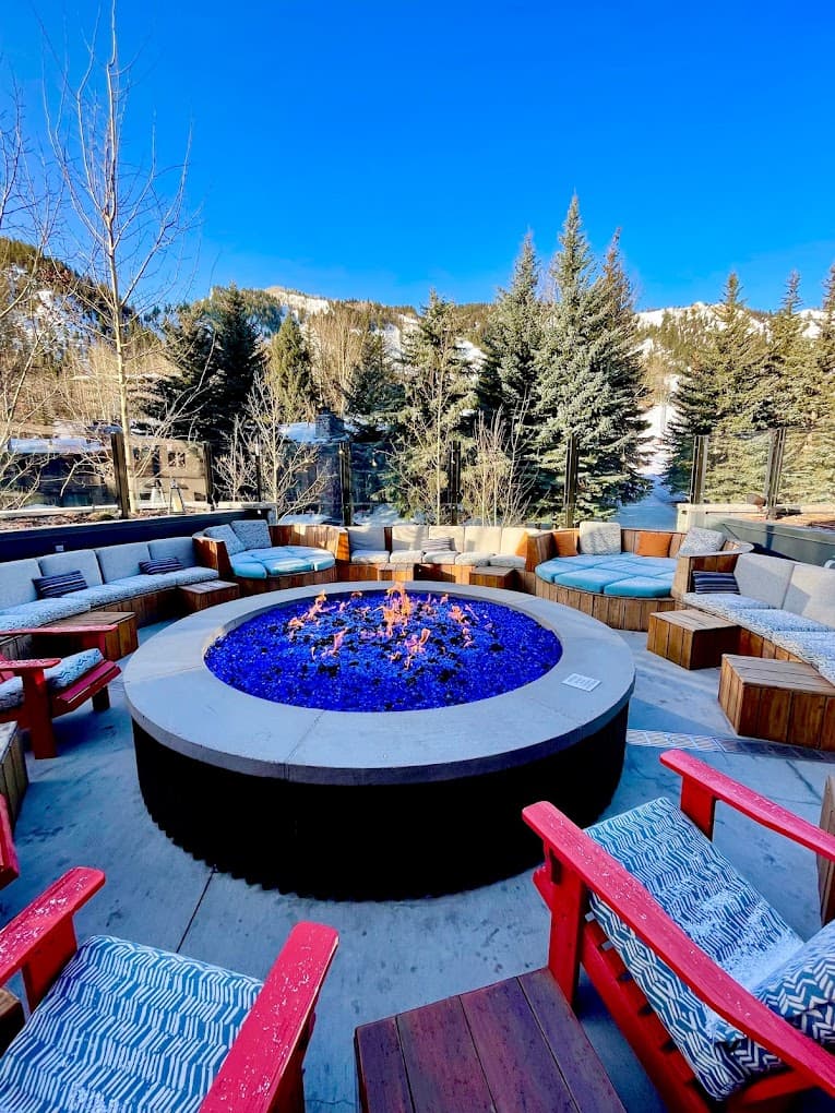 The fire pit at the W Hotel in Aspen features a roaring fire in the middle of rows of comfy seating near the trees on a clear day.