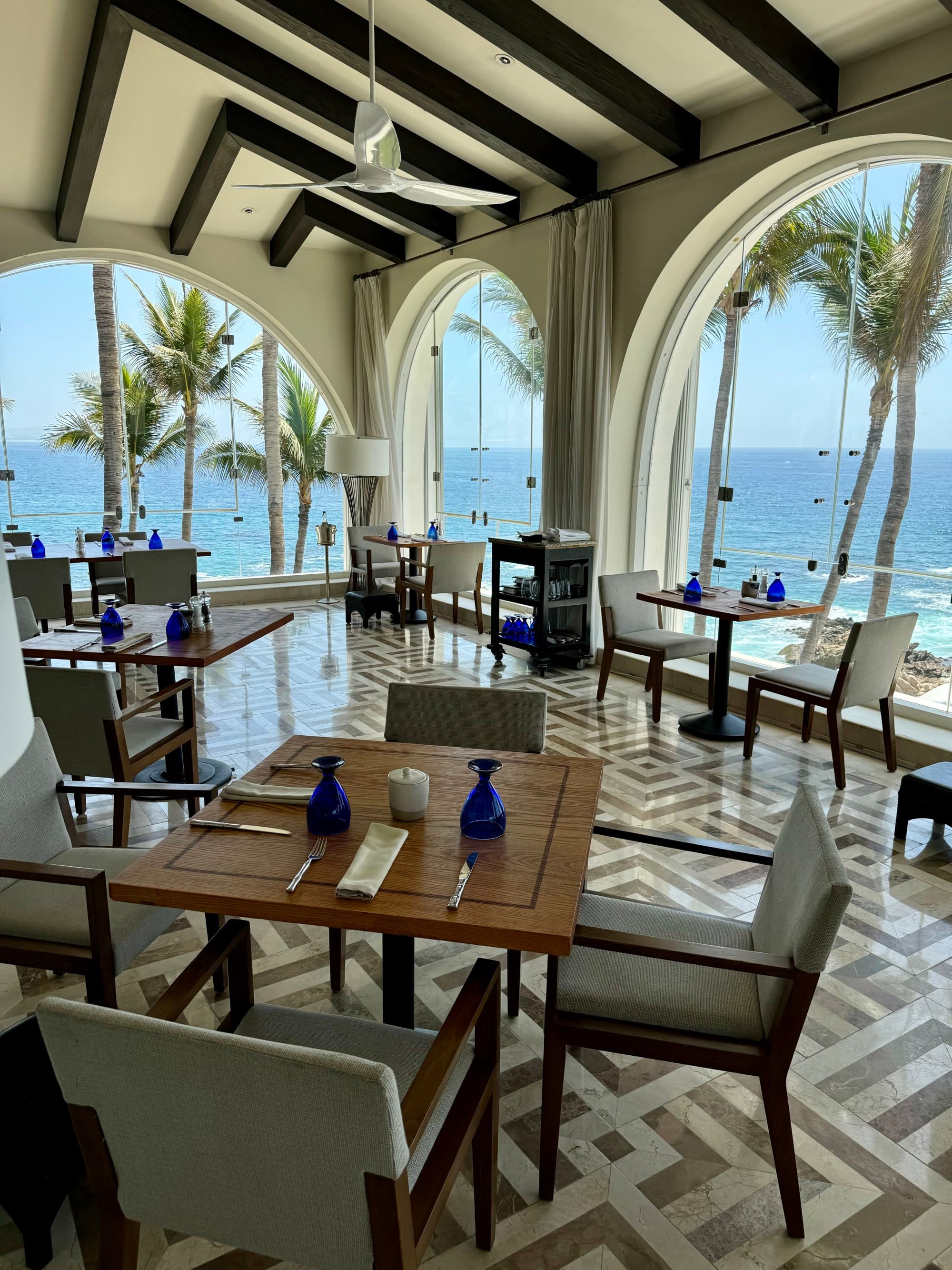 Outdoor dining area with wooden tables and chairs, open archways with views of the beach and gray and white geometric flooring in a Hotel in Baja California Sur
