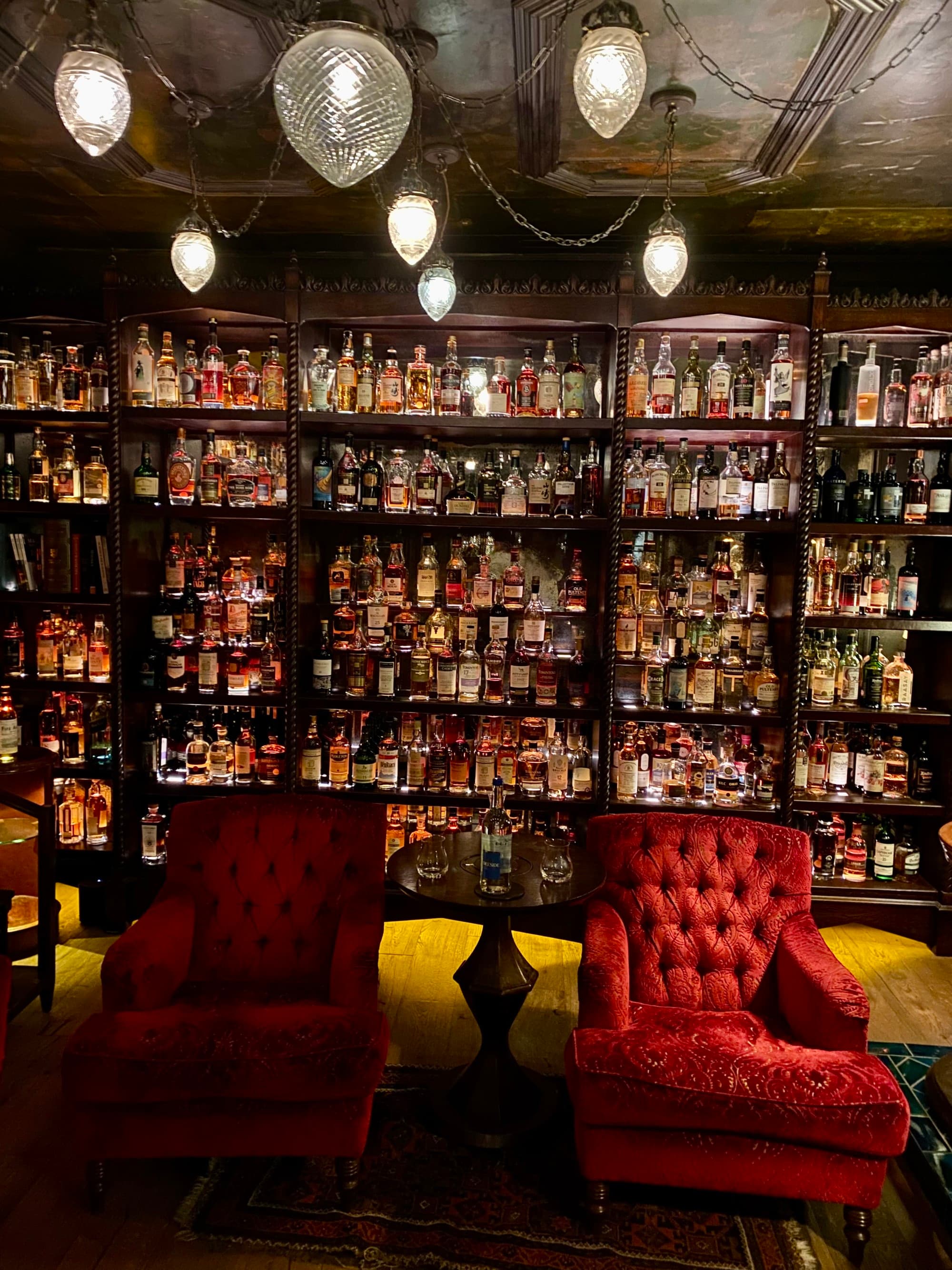 Shelves with hundreds of whiskey bottles in a dark room with velvet chairs.