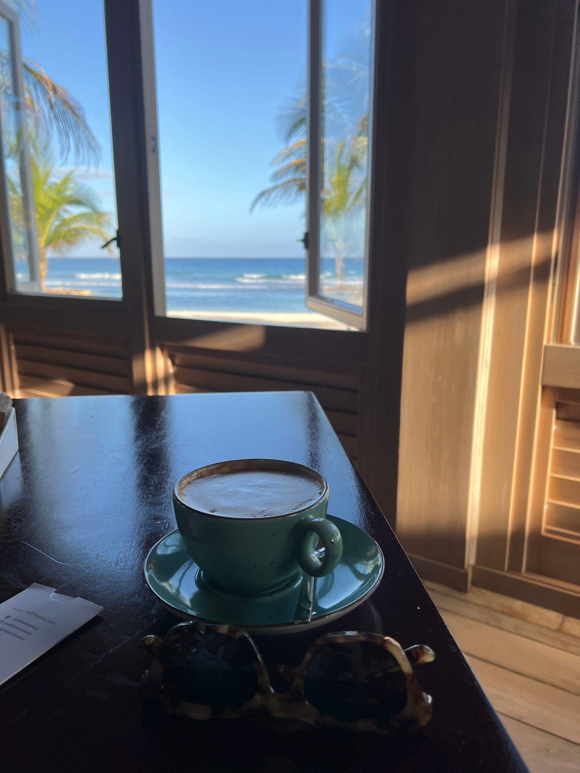 Coffee and sunglasses on a table with ocean visible through the window in the background.