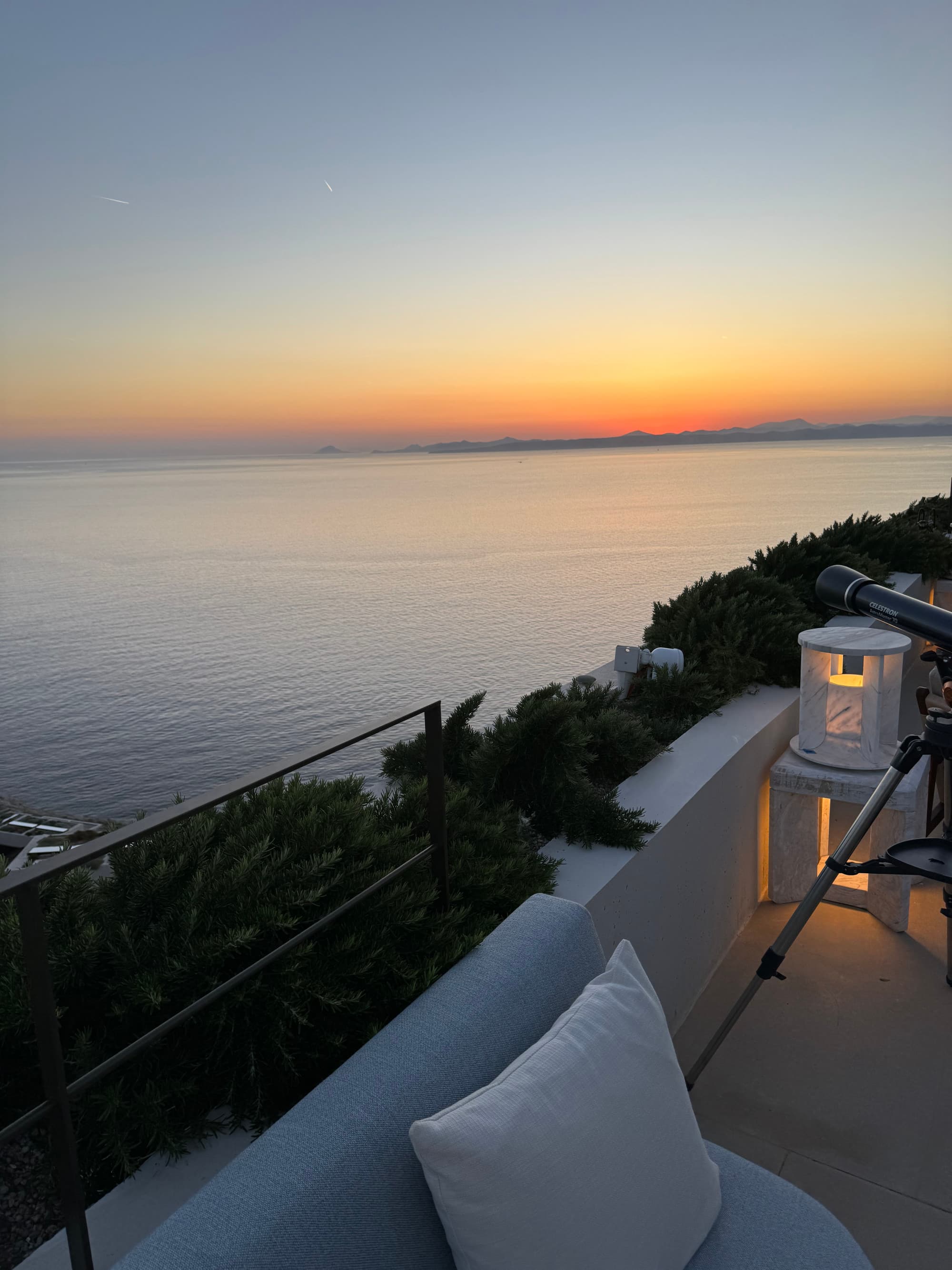 A patio with a couch and telescope overlooking the water at sunset.