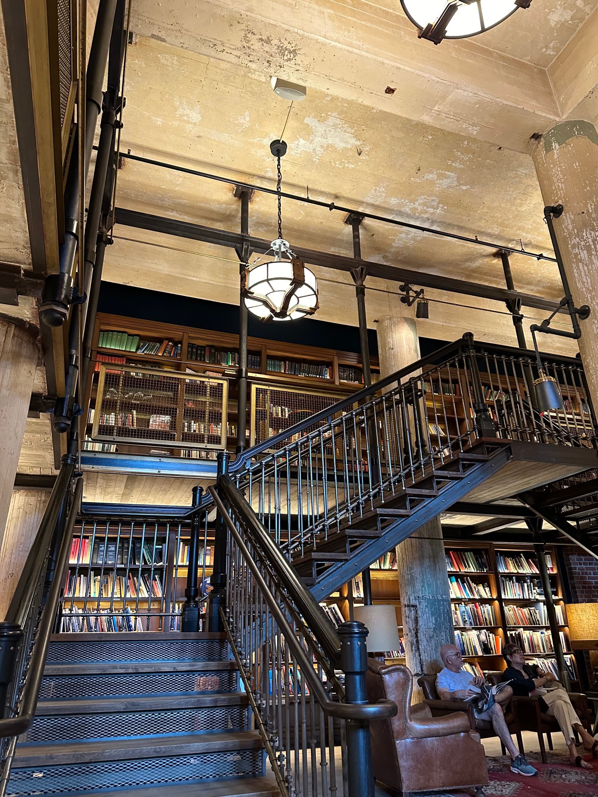 A library and sitting room featuring an iron staircase leading up to a balcony halfway up the bookcases.