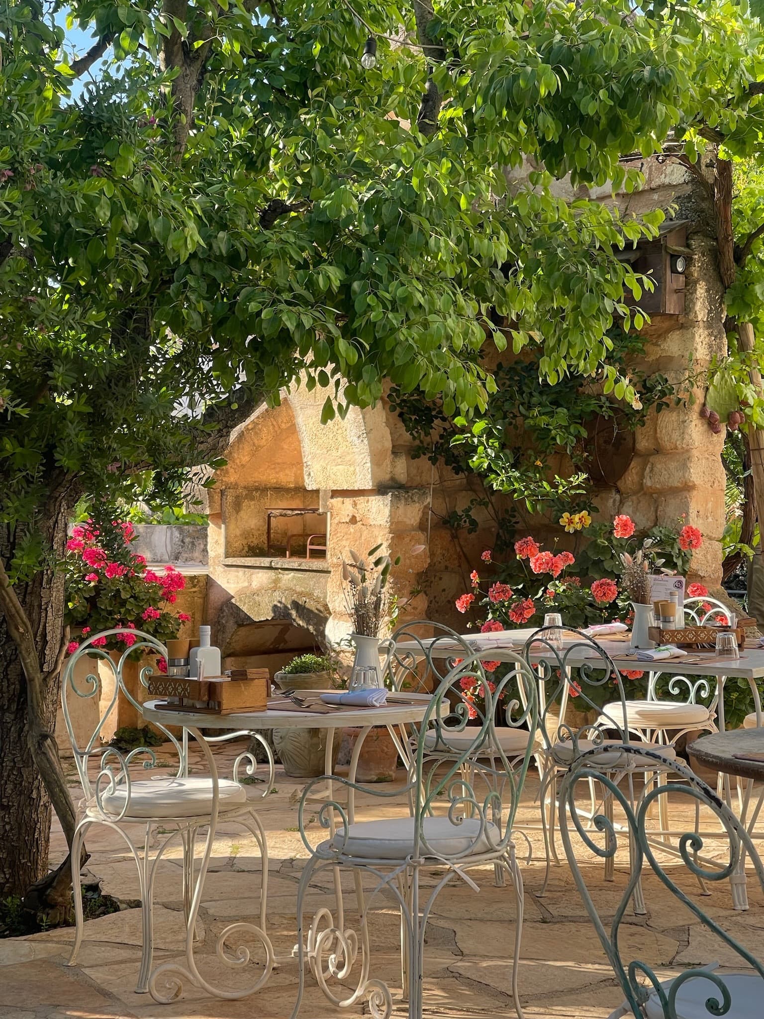 White, whimsical tables and chairs set in an outdoor courtyard, with pink flowers and green trees on a sunny day.
