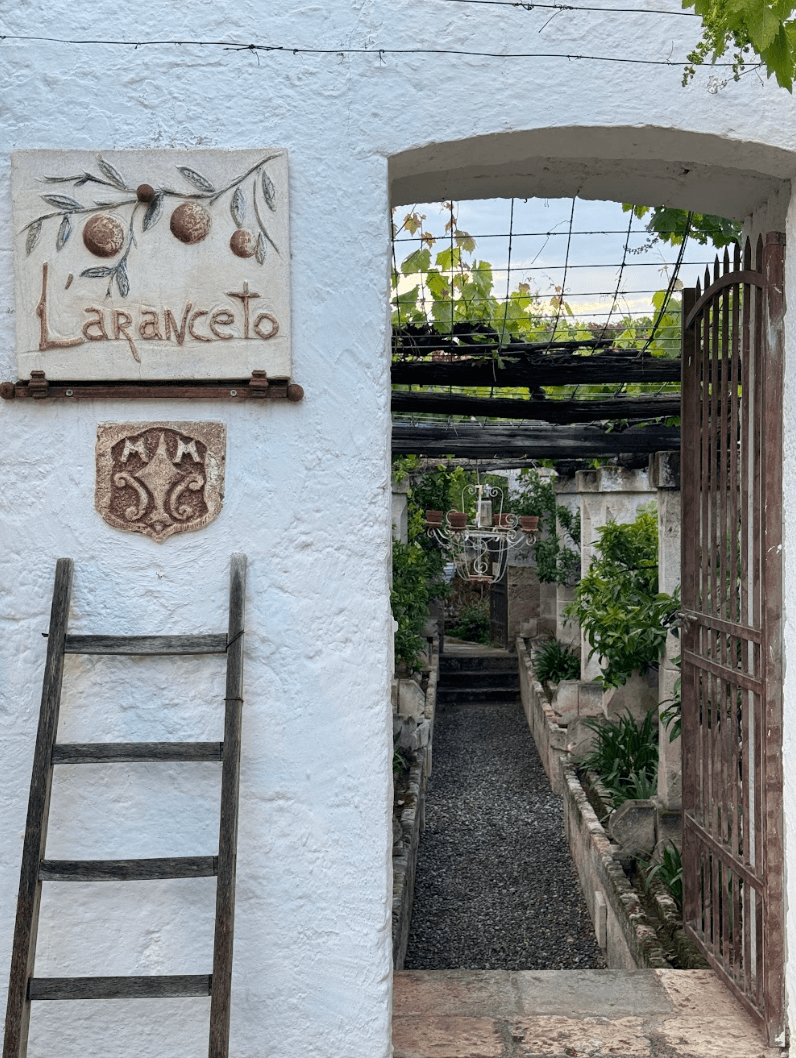A hand-painted sign hanging on a stucco wall, with a wooden ladder, and an open door revealing a garden pathway.