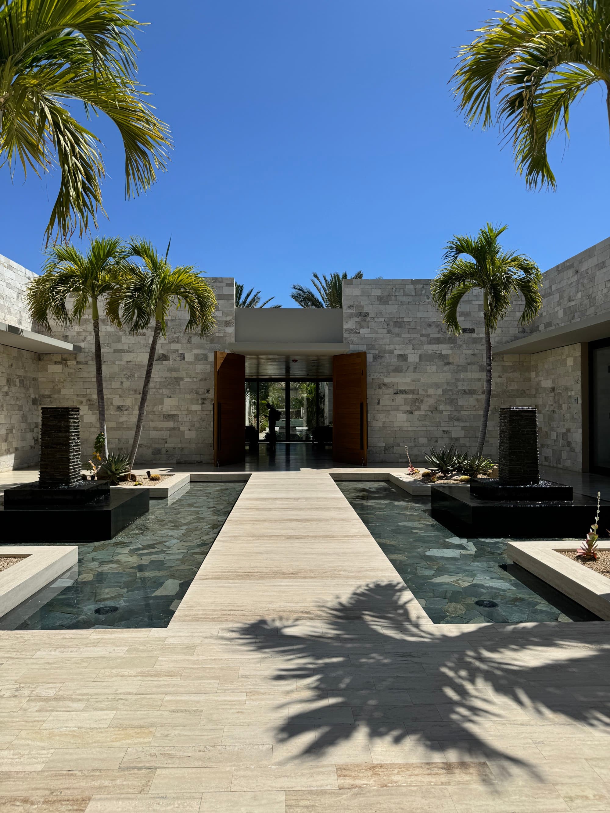 A view of the grounds of the resort, with modern-style fountains and palm trees on a sunny day.