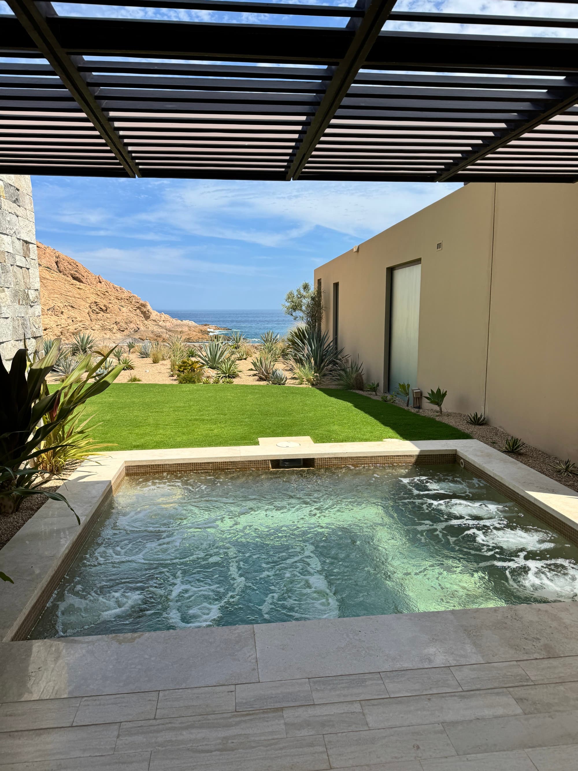 A sunny view of an outdoor hot tub, with running jets, leading to a grassy area, with the sea in the distance.