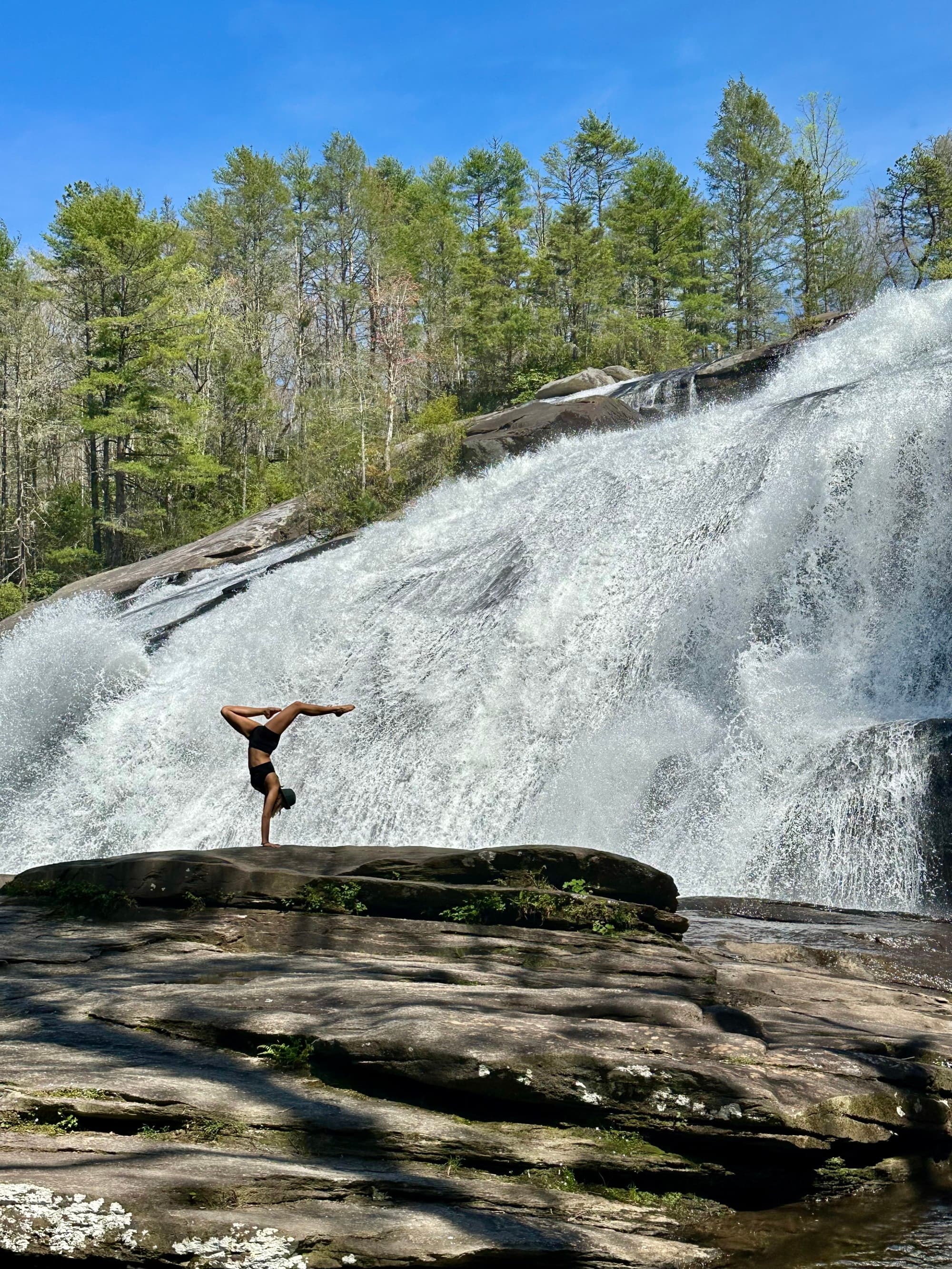 A young woman in a black outfit doing a handstand on a giant rock in front of a waterfall on a sunny day.