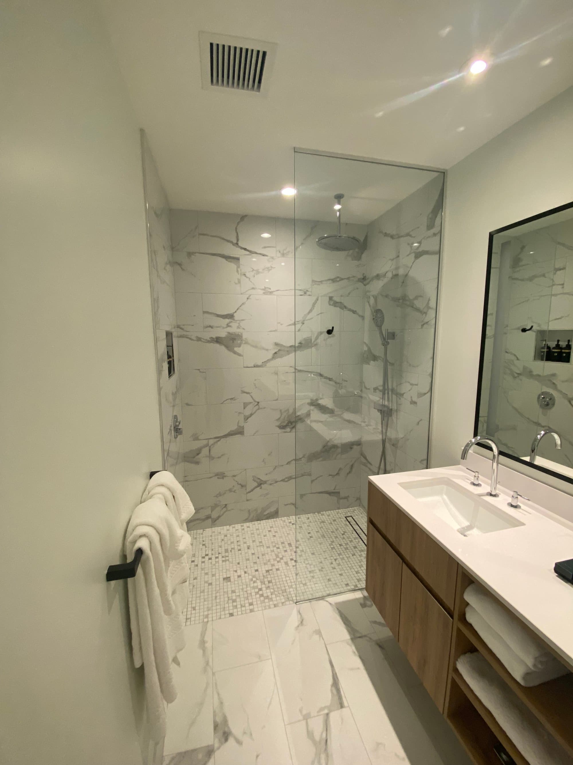 Inside the hotel bathroom, showcasing the shower stall, sink and towels, all in white and gray tones.