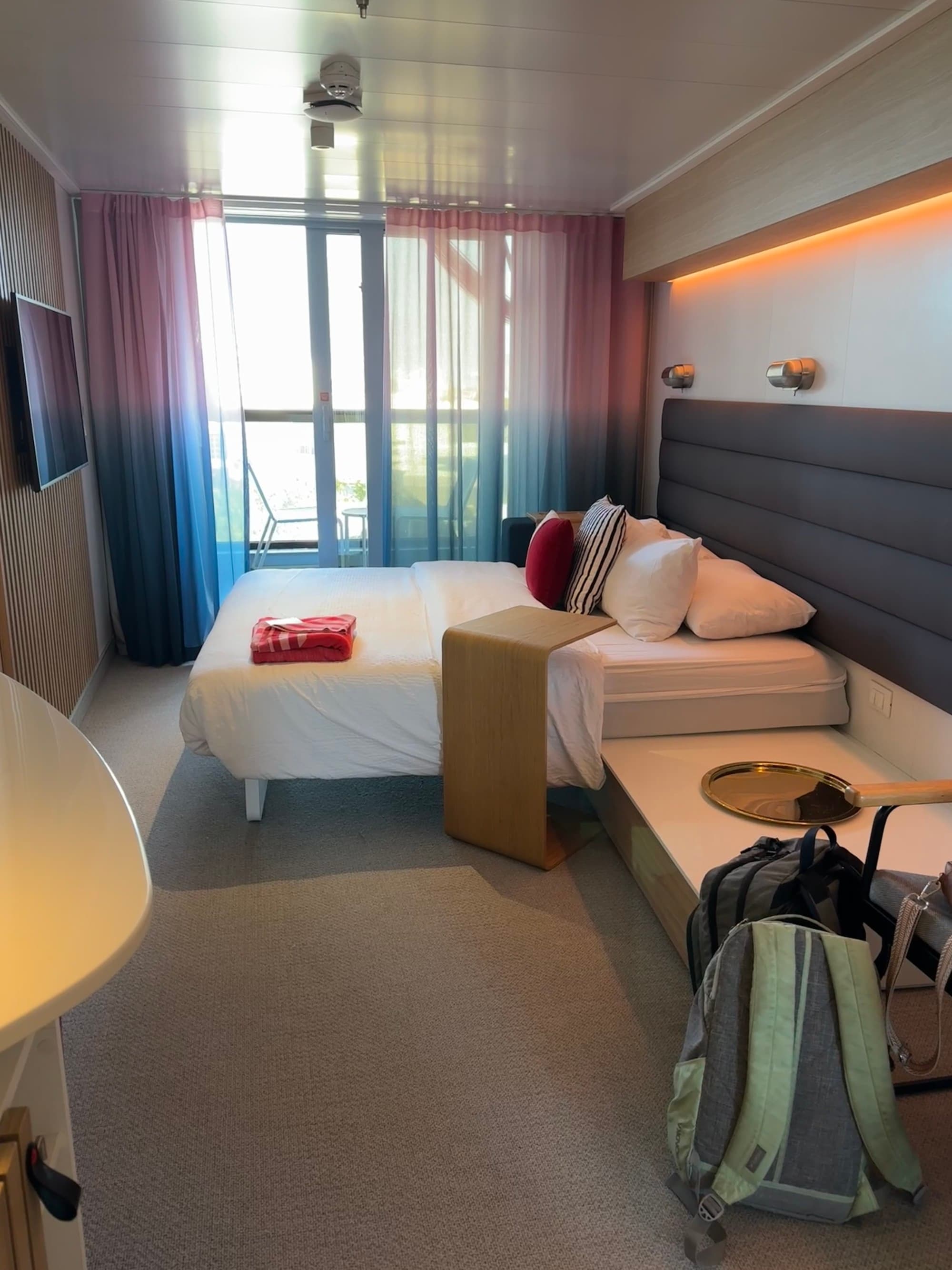 Inside the cabin with a master bed, table, television and glass doors leading to a sunny balcony.