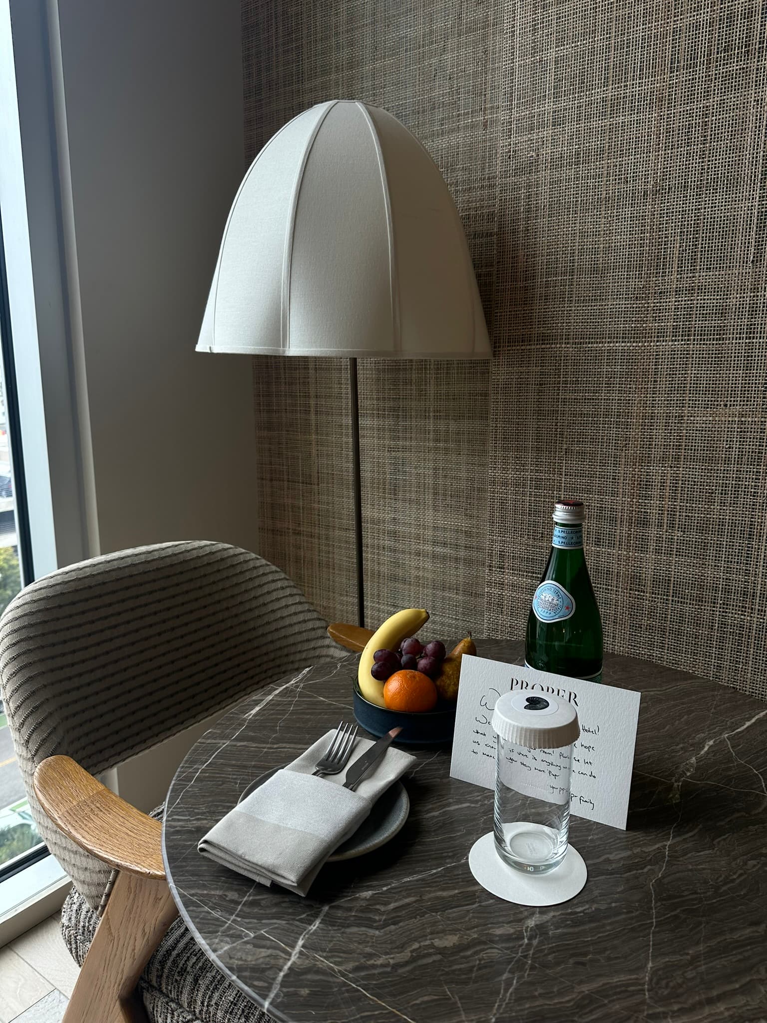 A soft desk chair, and round corner table topped with a note, fruit basket and bottle of sparkling water, inside of the hotel room.