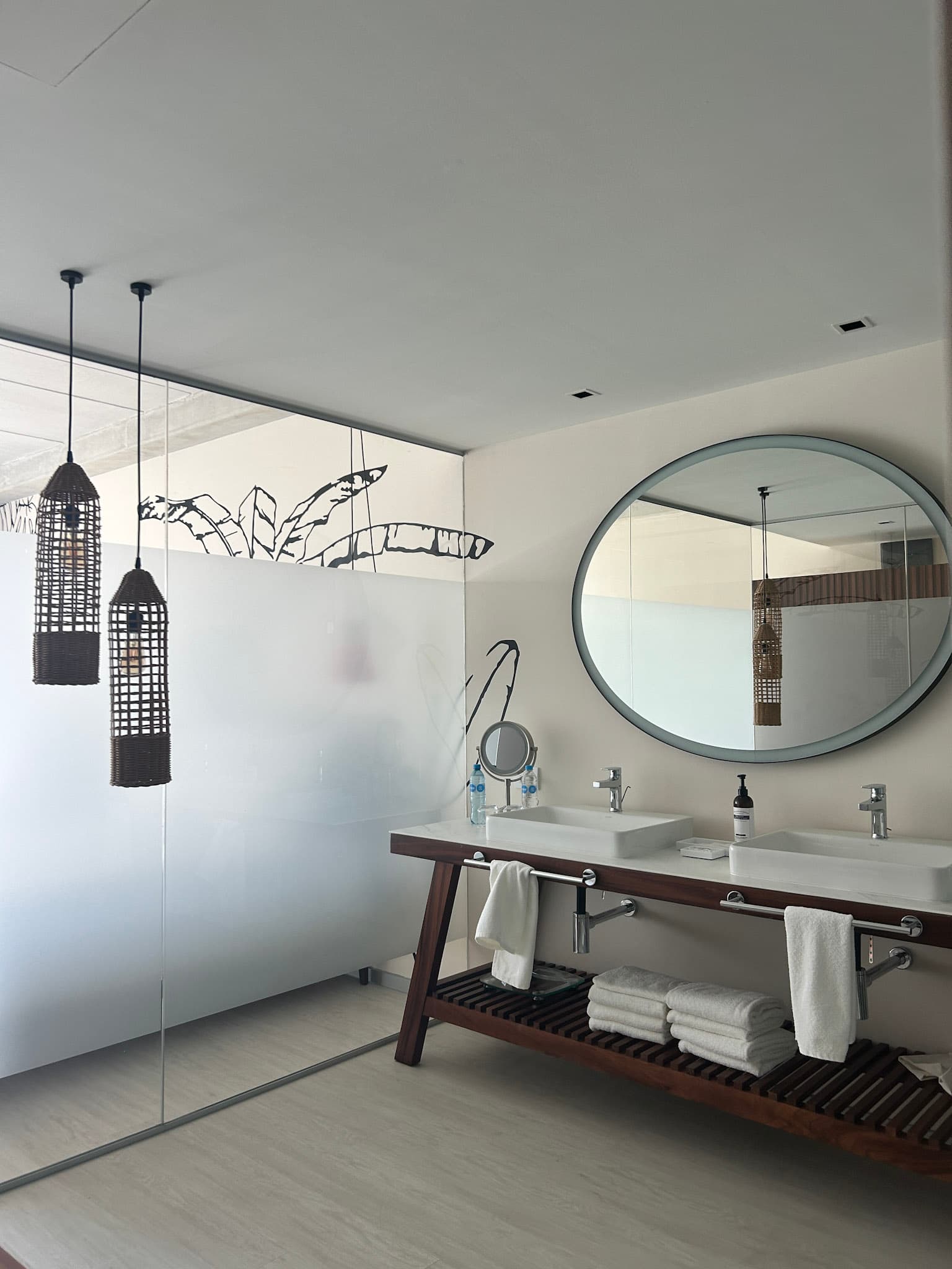 Inside the hotel room's bathroom, with a large, oval mirror, hanging light fixtures, double sinks and two stacks of folded, white towels.