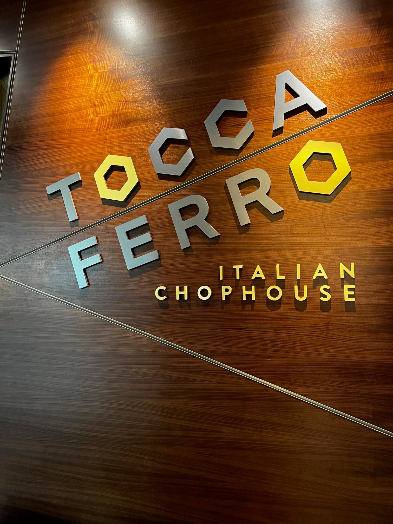 This image depicts a wooden wall with silver and yellow signage attached to it that reads "Tocca Ferro Italian Chophouse" beneath accent lighting.