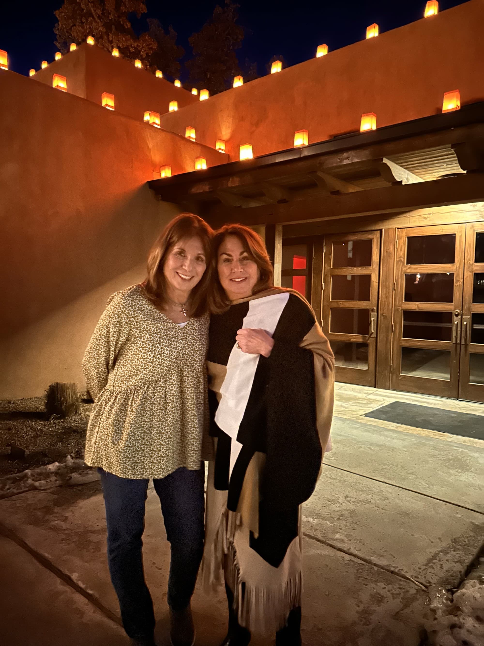 Two women with their arms wrapped around each other, in front of Bishops Lodge at night.