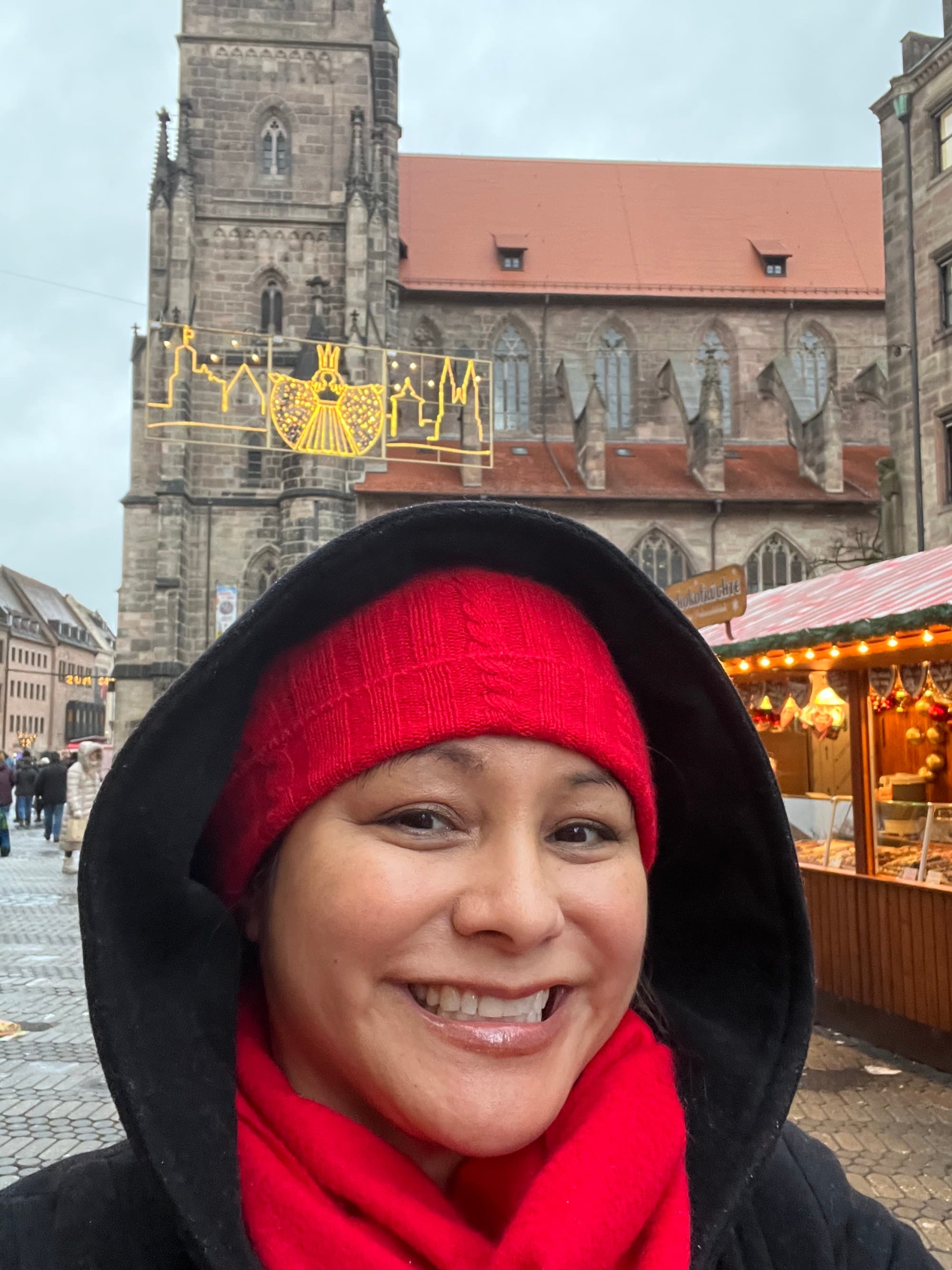 A person posing for a selfie outside while wearing a red knit hat and black hoodie. There is European architecture and holiday market huts in the background.