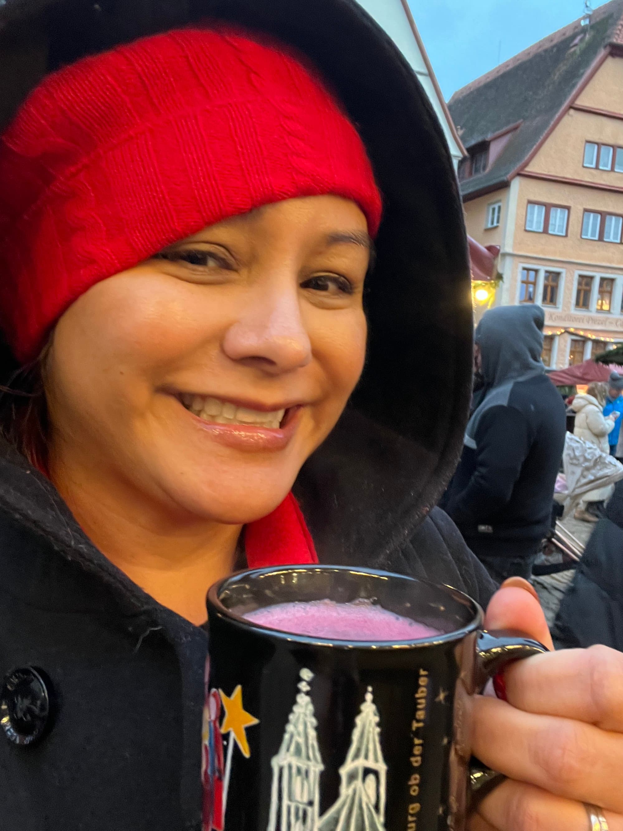 A person wearing a red knit hat and smiling while holding a mug outside with a purple liquid inside of it.
